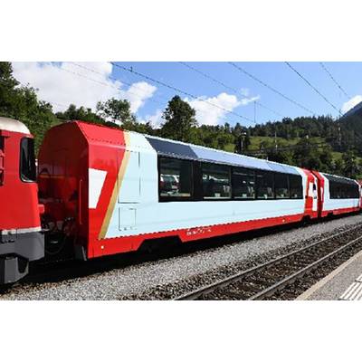 Rdeč in bel vlak vozi po tirnicah skozi zeleno, hribovito pokrajino pod modrim nebom z nekaj oblaki.