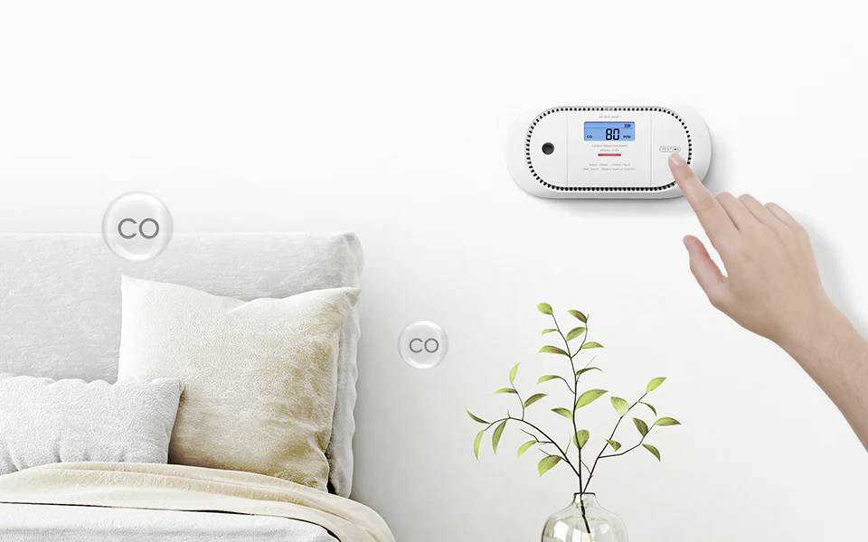 Roka nastavi digitalni detektor ogljikovega monoksida, ki prikazuje '80', v spalnici z posteljo in majhno rastlino, kar nakazuje na spremljanje kakovosti zraka.