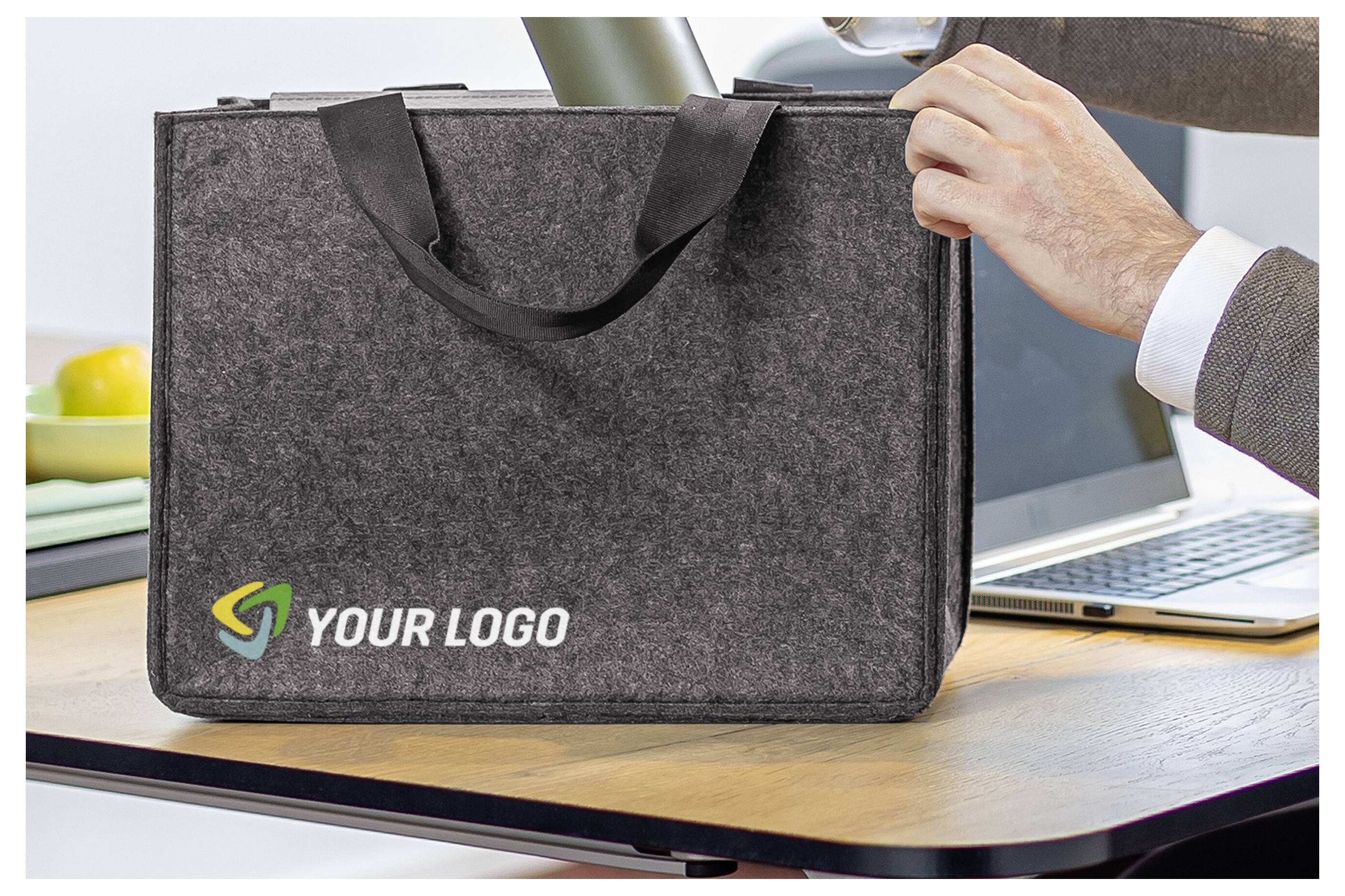 Roka, ki drži sivo filcno vrečko za nakupovanje s tiskom »YOUR LOGO« na pisalni mizi, v ozadju je prenosnik.