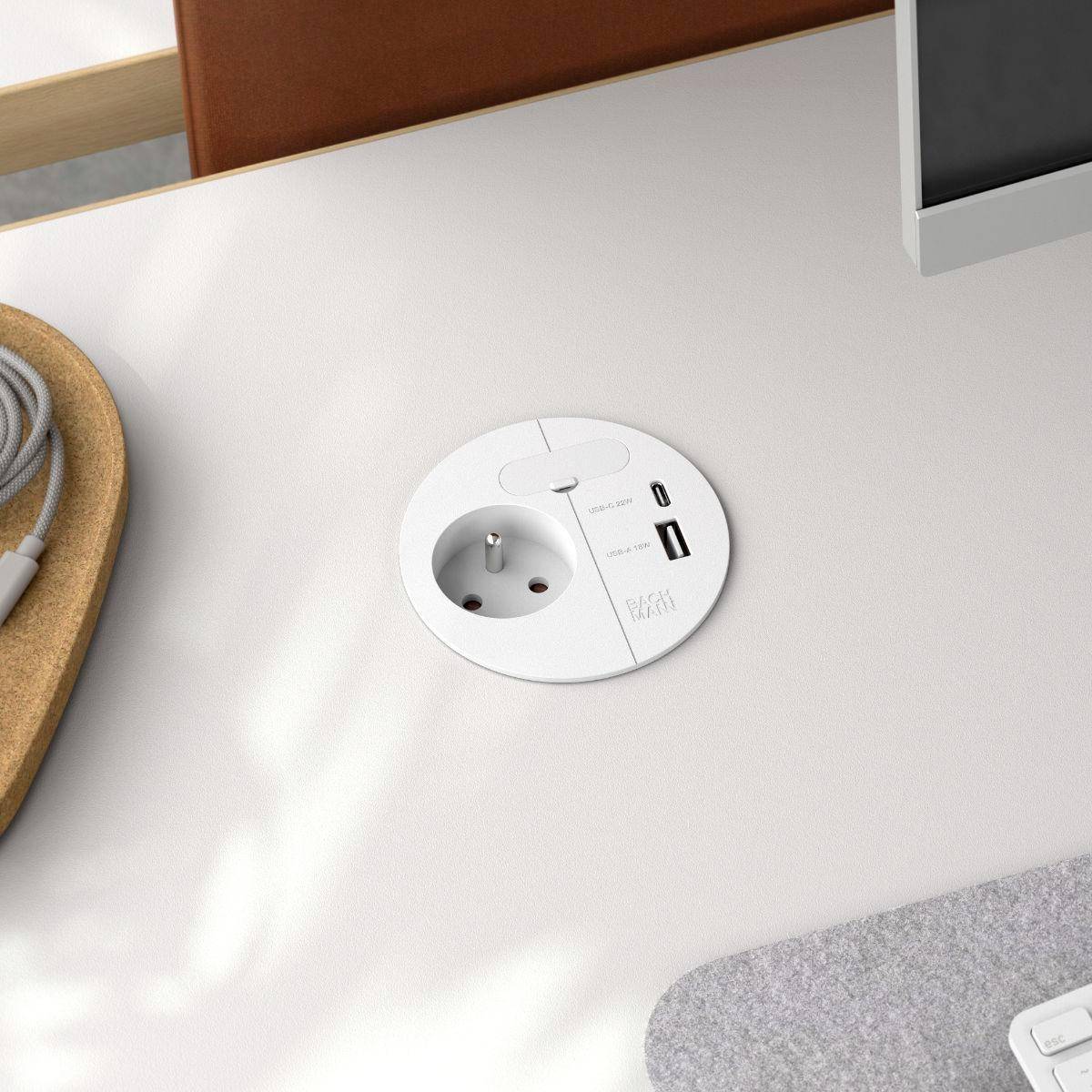 Vgrajena namizna okrogla električna vtičnica z USB vrati poleg zaslona računalnika, lesene površine mize in navite kable na levi strani.