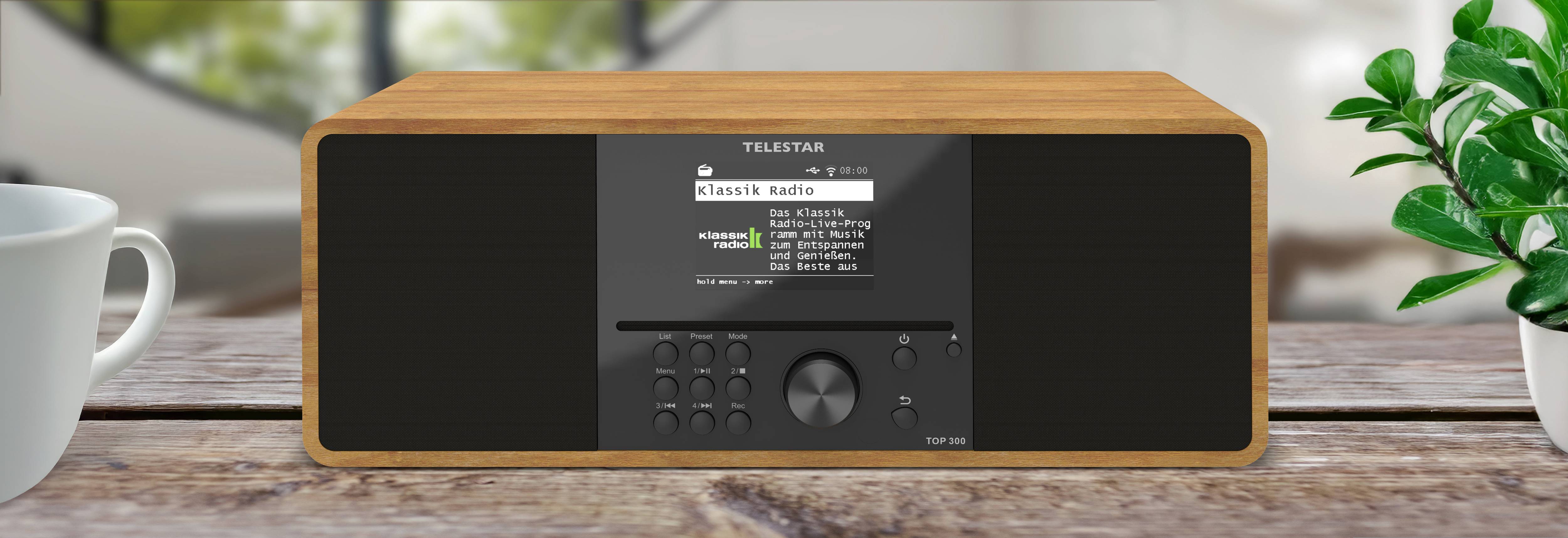 Pravokotni digitalni radio z lesenim okvirjem stoji na leseni mizi ter ima zaslon, kontrolne gumbe in številčnico.