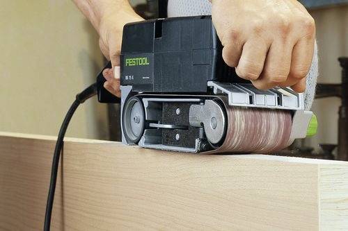 Oseba uporablja električni tračni brusilnik za glajenje lesene površine, pri čemer se osredotoča na natančnost in enakomerno obdelavo.