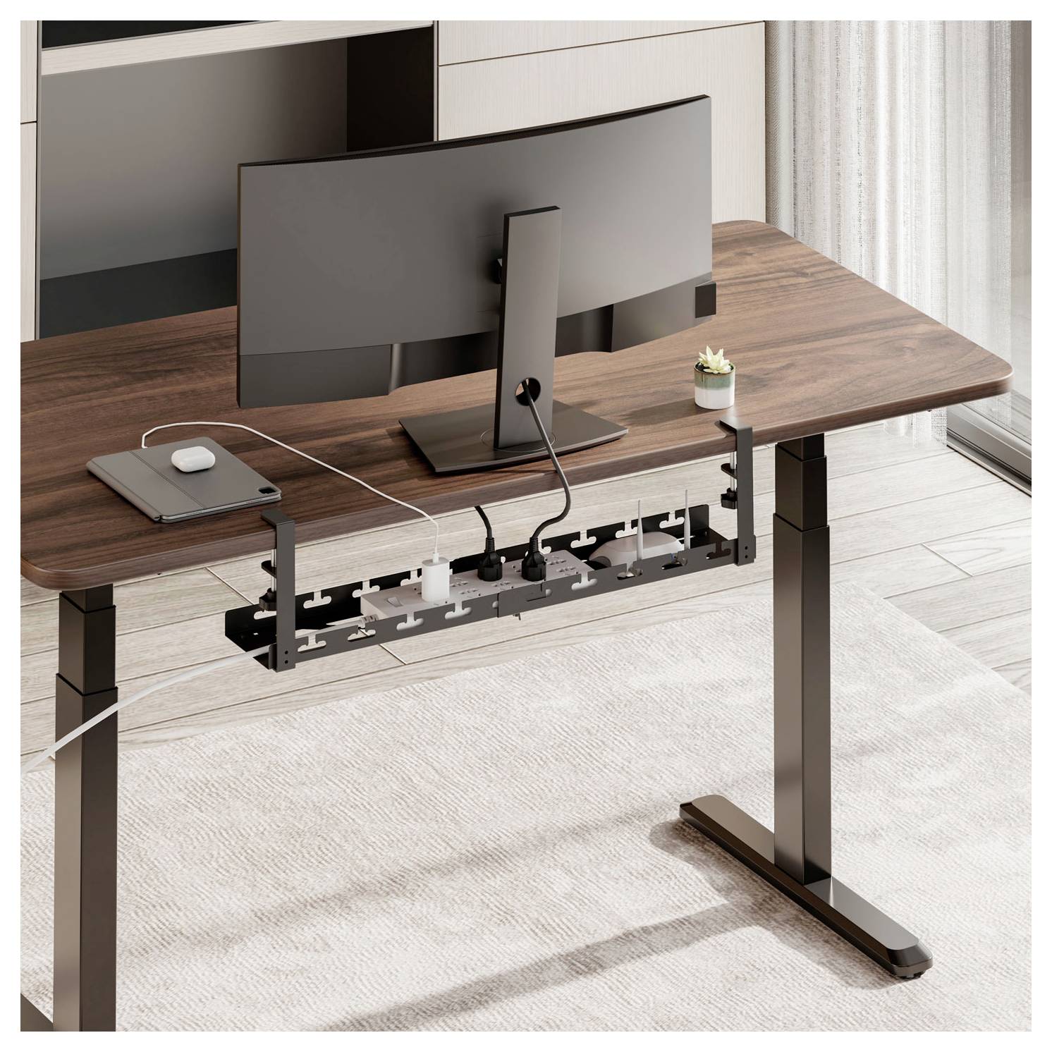 Moderna pisarna z leseno mizo, na kateri je črn monitor, prenosni računalnik in razdelilec za elektriko spodaj. Majhna rastlina dodaja dotik zelenja.