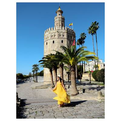Ženska v lahnem rumeno-rumenem oblačilu hodi blizu Torre del Oro, v ozadju pa so palme in jasno modro nebo.