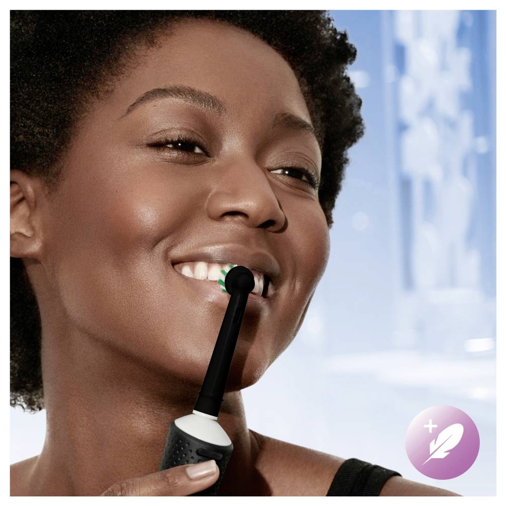 Oseba, ki se nasmehuje med uporabo električne zobne ščetke, z zamegljenim ozadjem in ikono peresa, ki simbolizira nežno čiščenje.