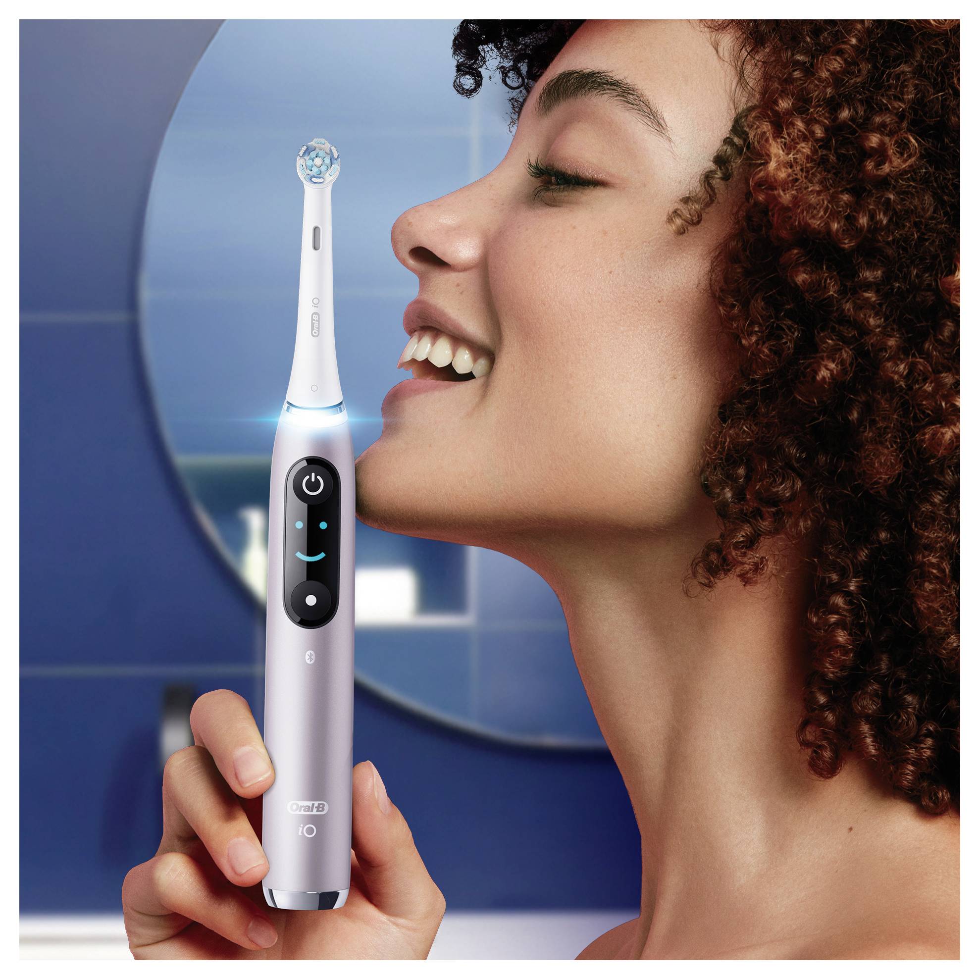 Oseba, ki se nasmihlja med tem, ko drži električno zobno ščetko blizu obraza, demonstrira njeno uporabo in poudarja skrb za zobno nego.