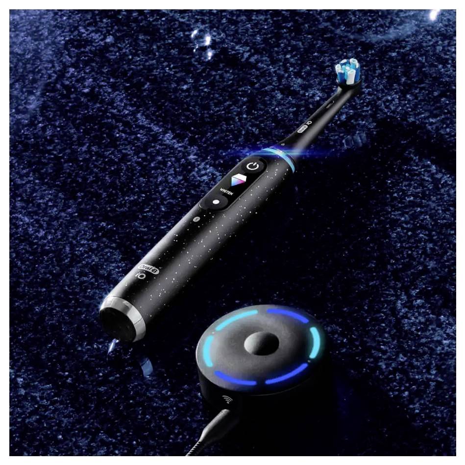 Električna zobna ščetka z osvetljenimi gumbi počiva na sijajni črni površini poleg krožne polnilne postaje.