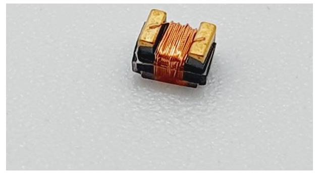 Majhna elektronska komponenta z bakrenimi žičnatimi tuljavami, verjetno induktor, postavljena na belo podlago.