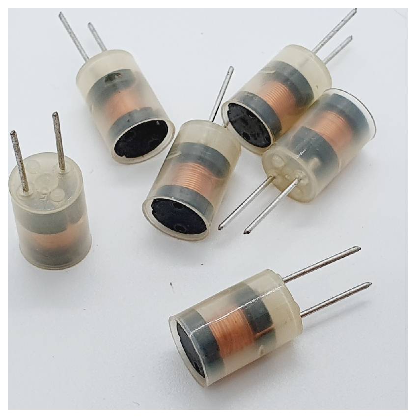 Šest majhnih elektronskih induktorjev z oranžnimi bakrenimi tuljavami, nameščenih v prosojnih valjih, postavljenih na beli površini.