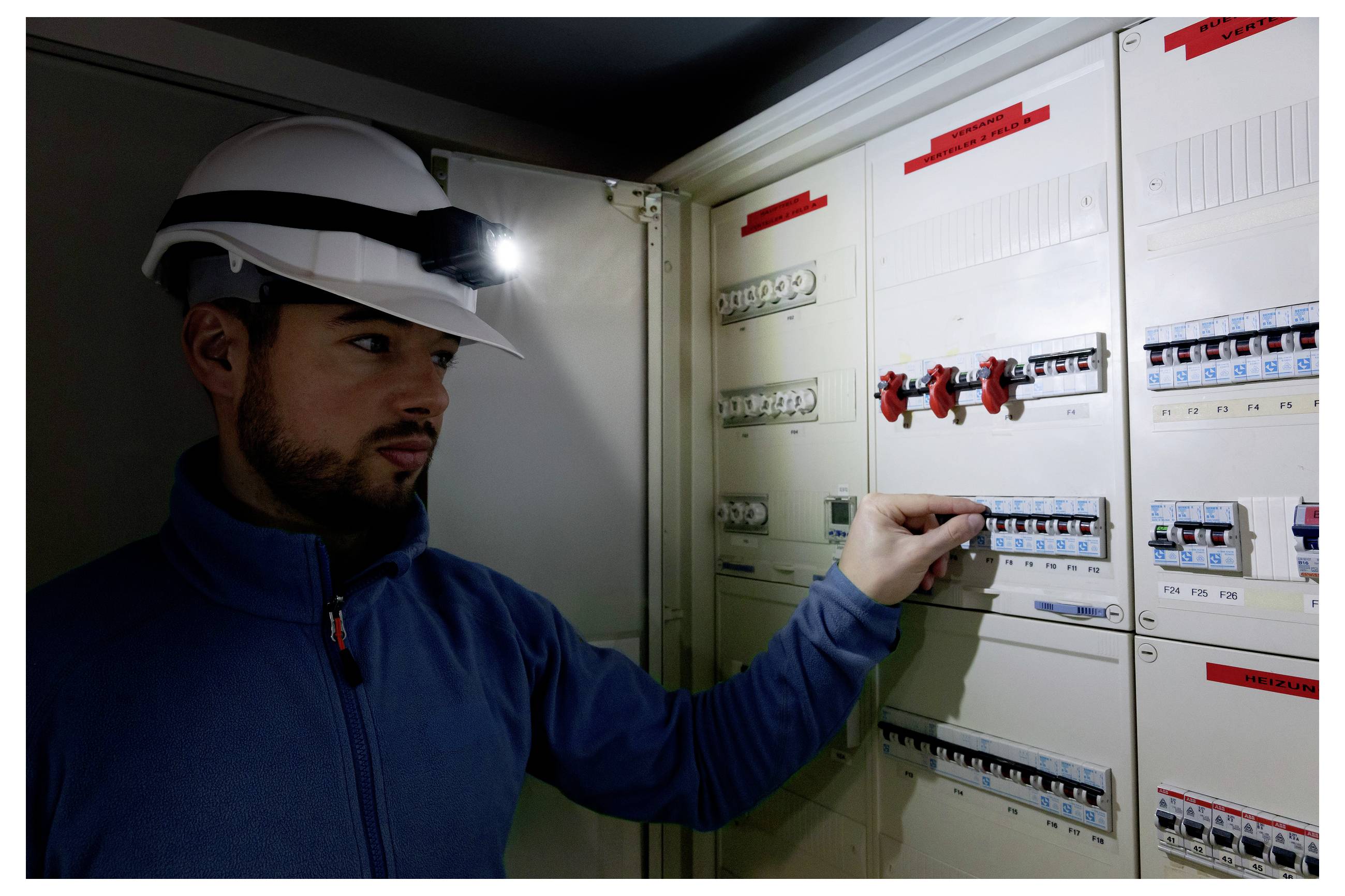 Električar v čeladi z naglavno svetilko pregleduje električno tablо, pri čemer preverja varnostne odklopnike v slabo osvetljeni sobi.