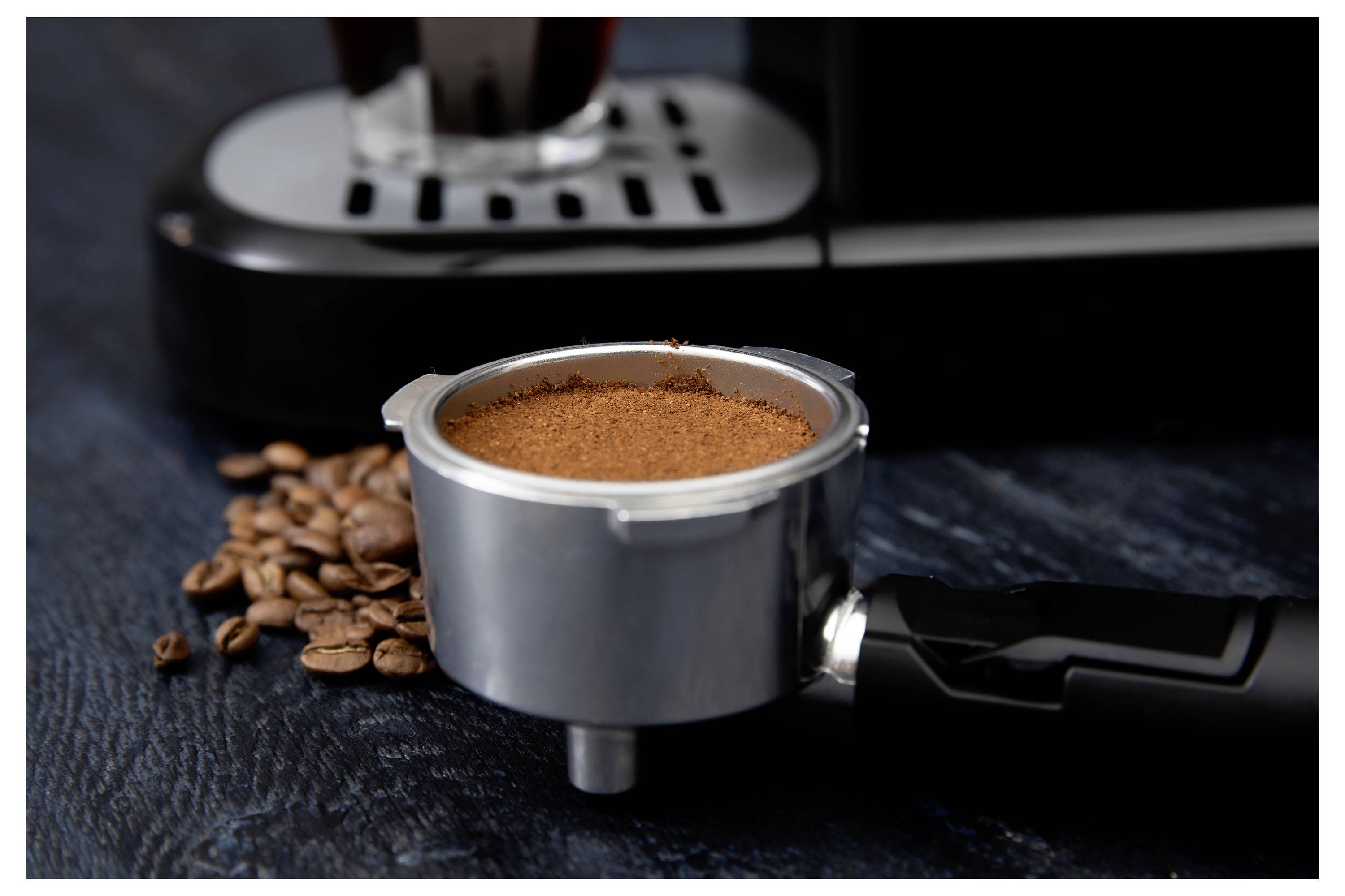 Portafilter, napolnjen z mleto kavo, poleg kavnih zrn, postavljen pred espresso aparatom na temnem ozadju.