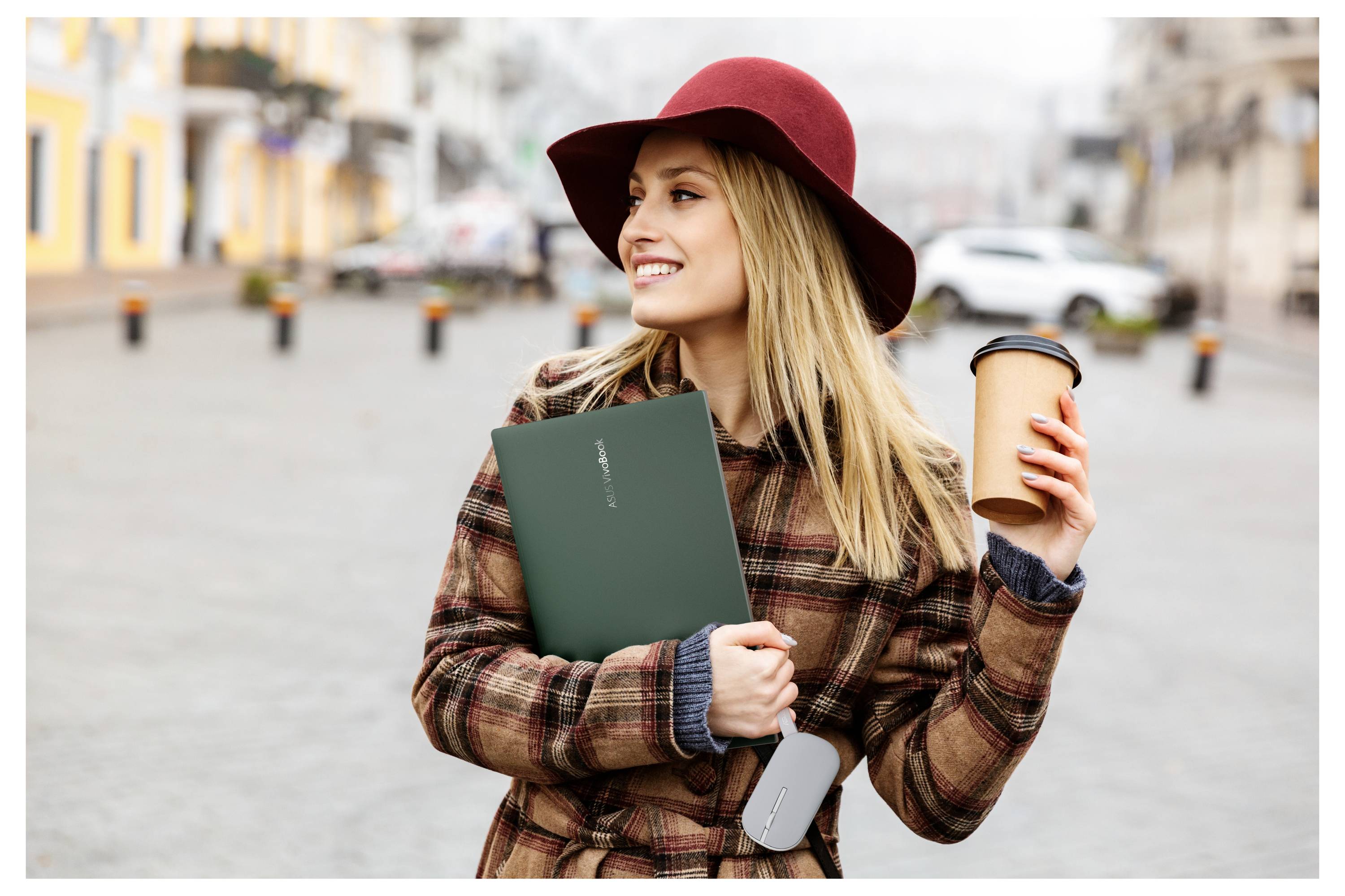 Ženska v karirasti plašč in rdeči klobuk stoji na prostem, drži skodelico kave in zeleno mapo, smeji se, z zabrisanim urbanim ozadjem.
