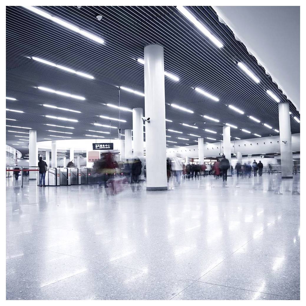 Prometna, široka podzemna postaja z visokimi stropi in fluorescenčno razsvetljavo, kjer se zamegleni potniki premikajo skozi prehode.