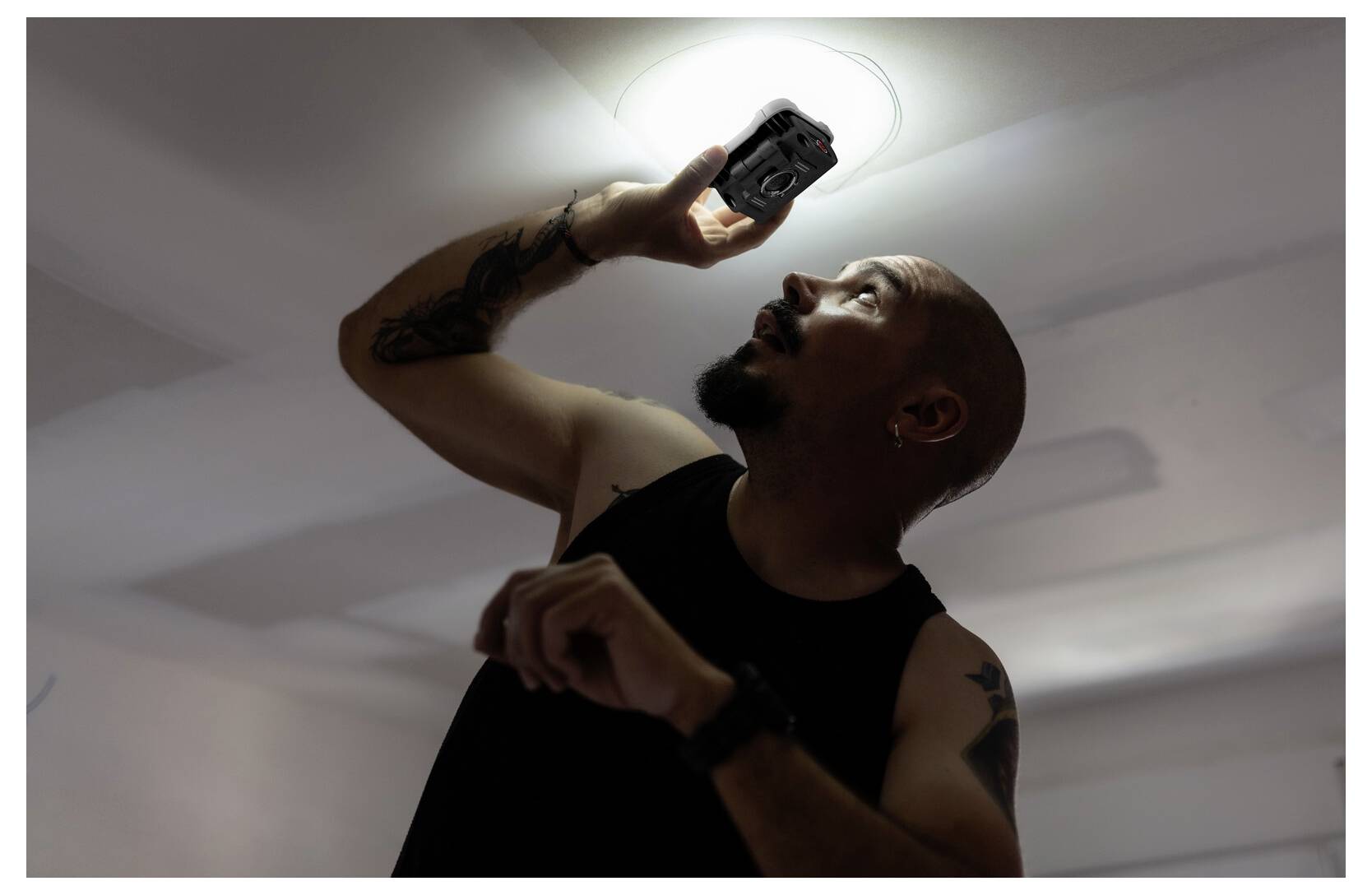 Oseba stoji na lestvi, drži fotoaparat visoko proti osvetljenemu strobnemu svetilu ter pregleduje ali fotografira nekaj zgoraj.