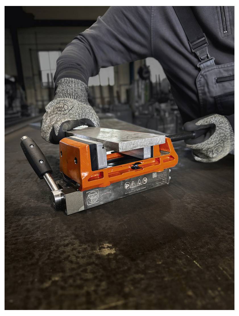 Delavec na velikem stolu v industrijski nastavitvi uporablja oranžno kovinsko brusiće, pri čemer nosi rokavice in temno predpasnik.