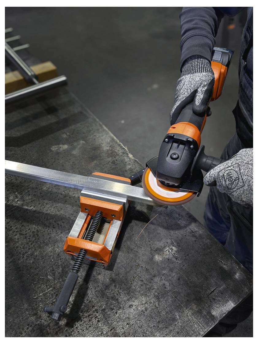 Oseba v zaščitnih rokavicah z oranžno in črno kotno brusilko reže kovina, pritrjeno v vijak na delovni mizi.