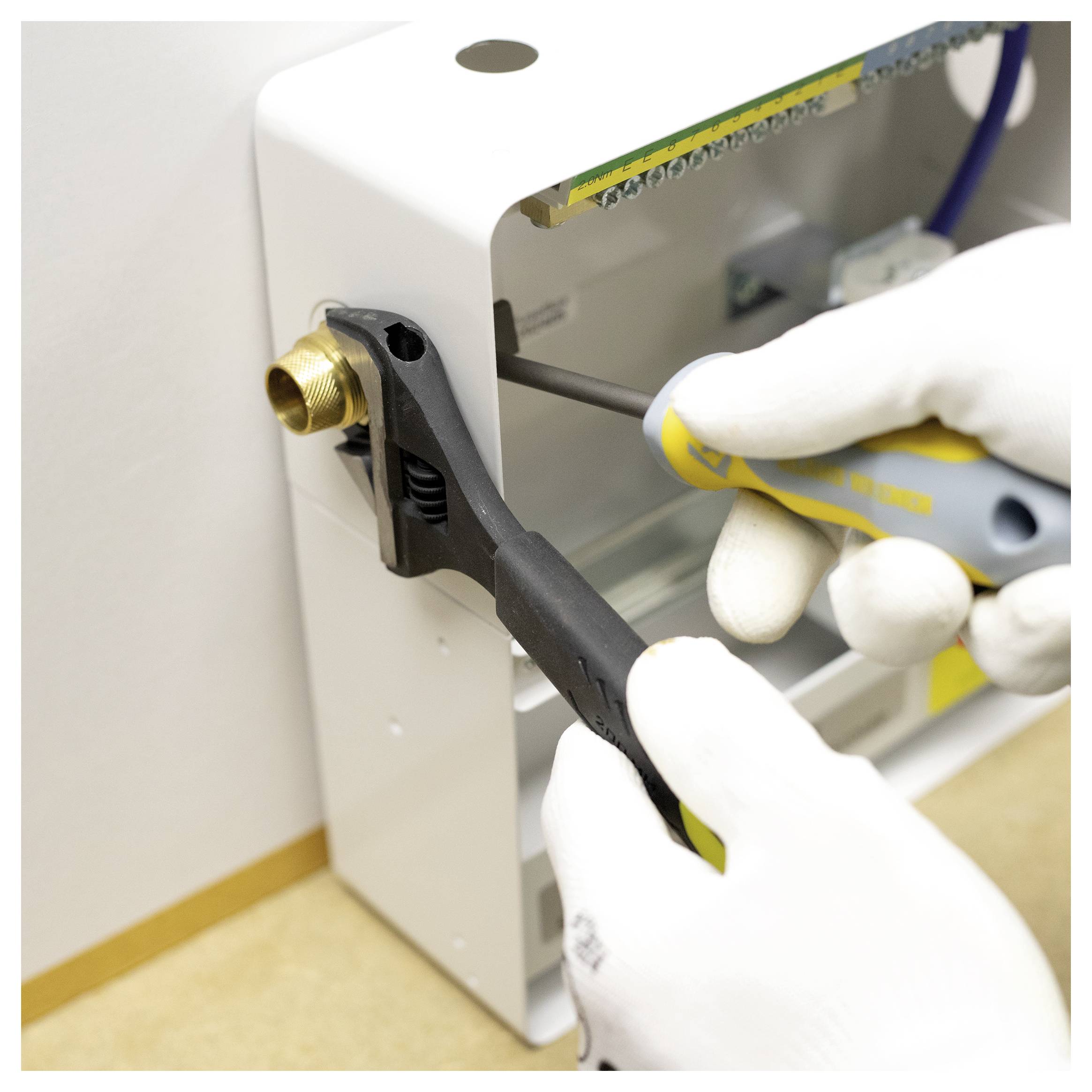 Oseba v rokavicah uporablja ključ za nastavljanje medeninastega matica na beli industrijski napravi, pri čemer prikazuje postopek vzdrževanja.