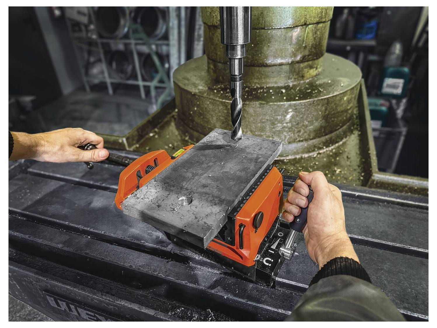 Dve osebi upravljata stiskalnico, pri čemer se osredotočita na poravnavo kovinskega kosa pod svedrom, kar prikazuje timsko delo in natančnost v delavnici.