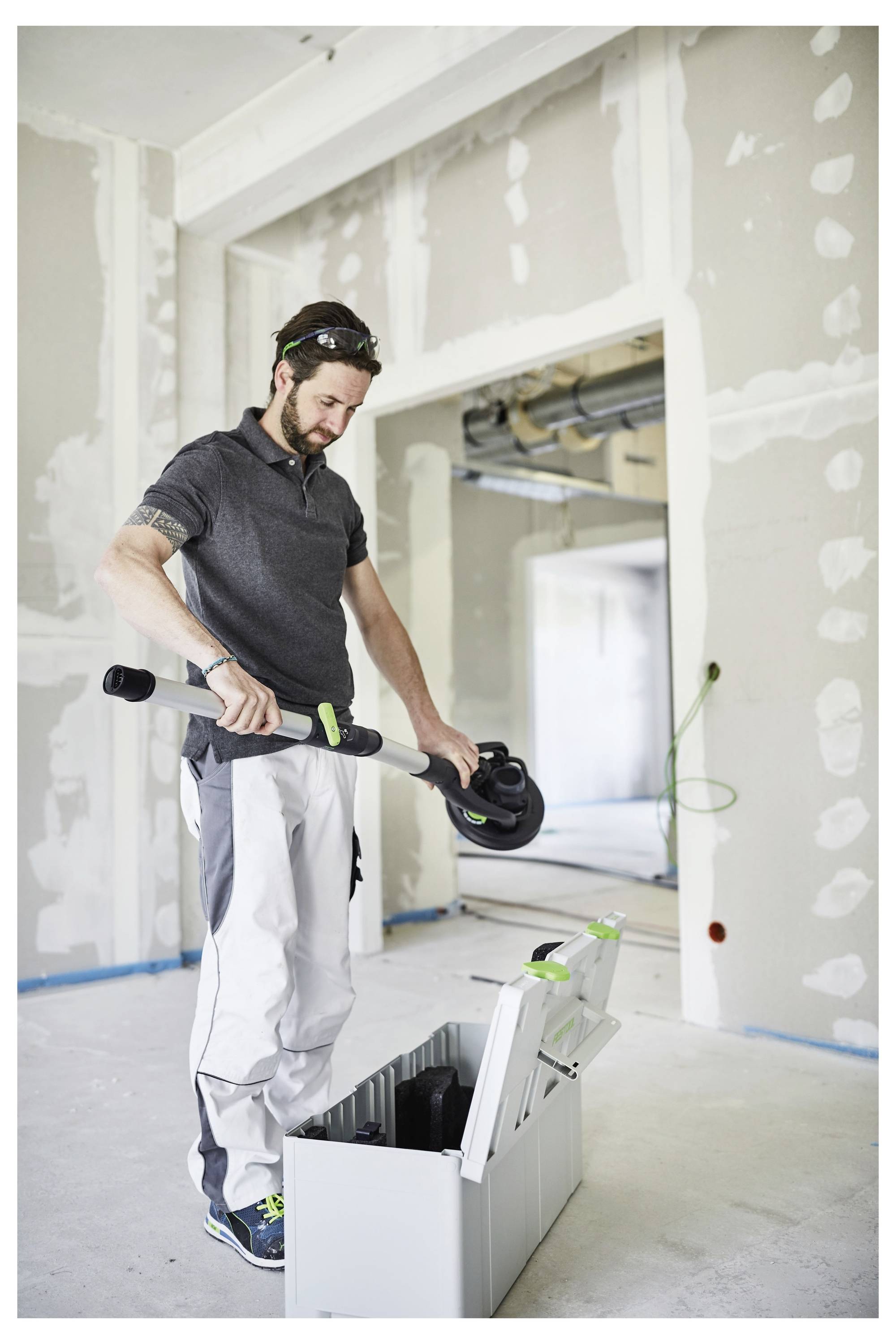 Oseba stoji v zaprtih prostorih in montira ali demontira orodje, verjetno za gradnjo ali prenovo, v ozadju pa so svetlostne stene.