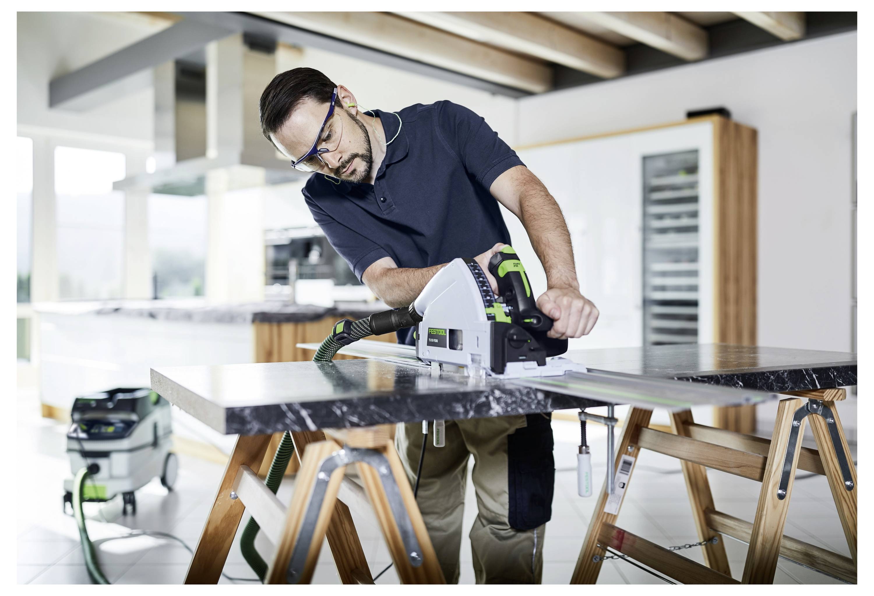 Moški s varnostnimi očali upravlja krožno žago na marmornati plošči v sodobni kuhinjski postavitvi. Je osredotočen in orodje rokovati z skrbnostjo.