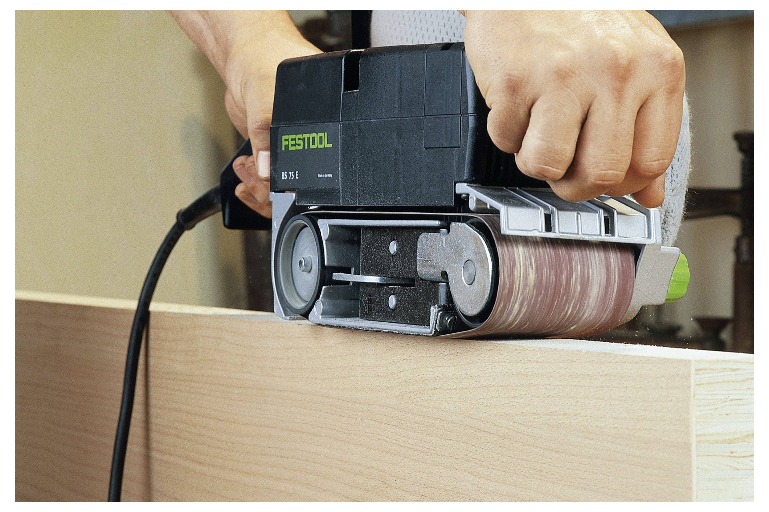 Oseba uporablja električni brusilnik na robu lesene deske in gladka površino. Brusilnik ima oznako »FESTOOL«.