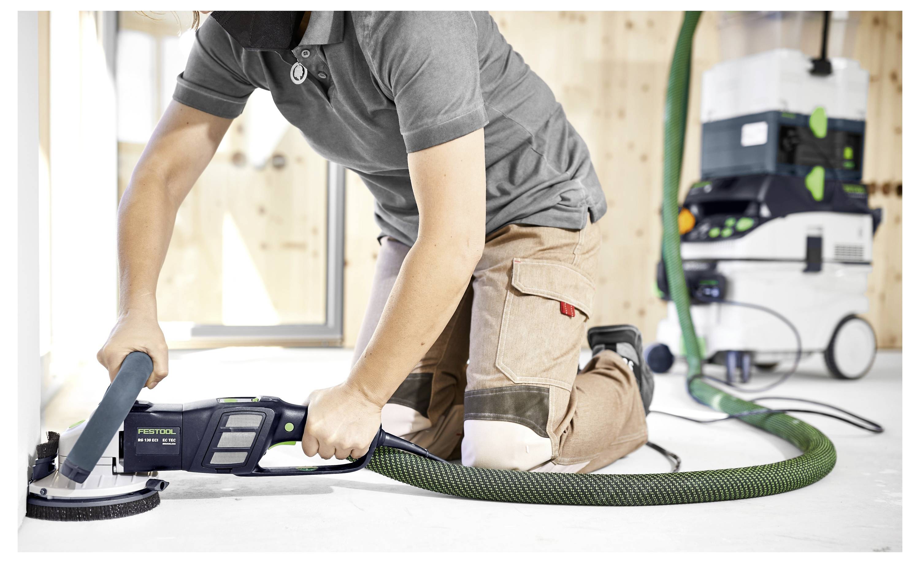 Oseba kleči na tleh in uporablja električni brusilnik s priklučenim zelenim cevjem na sesalnik za prah v delavnici za obdelavo lesa.