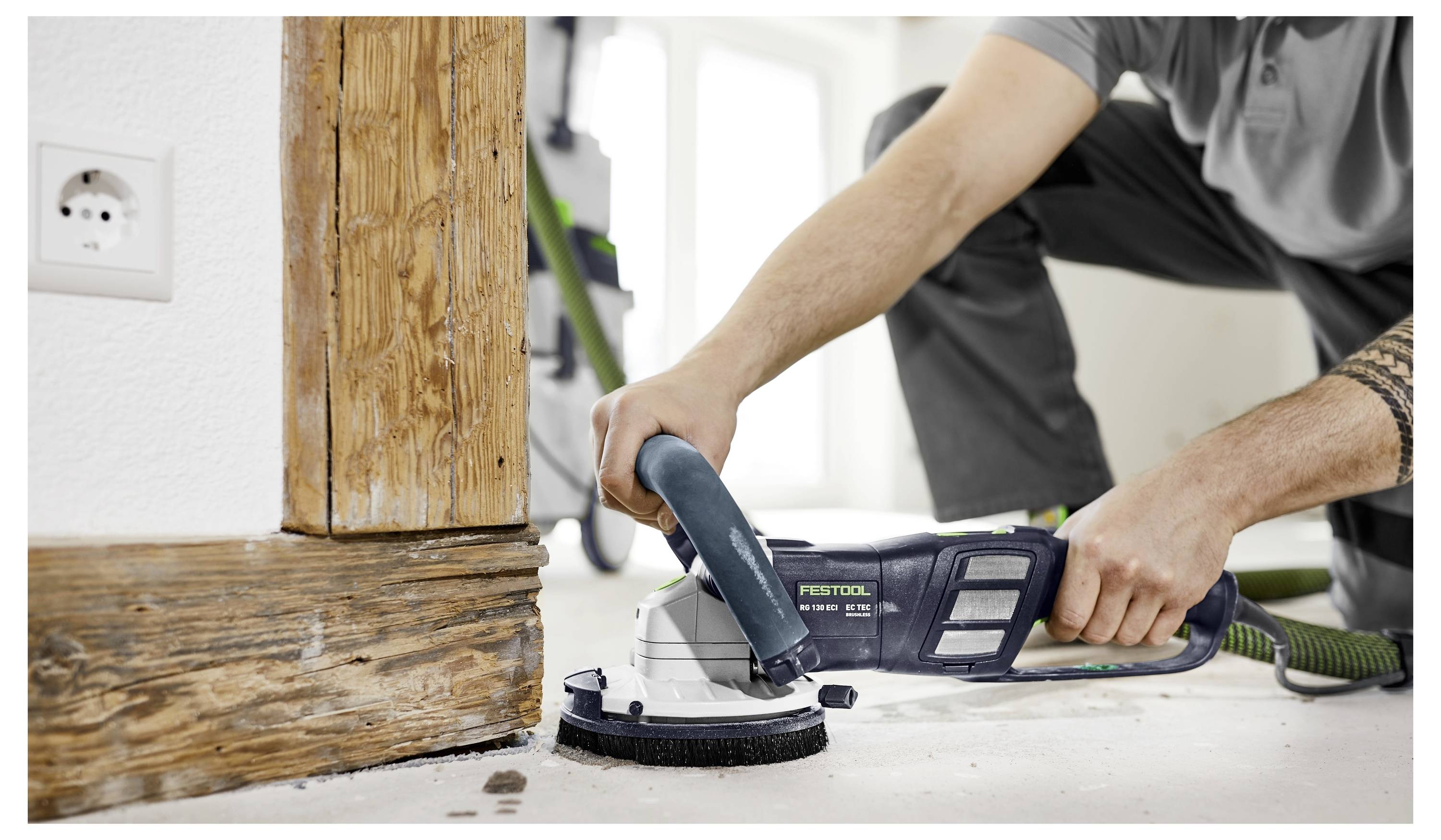 Oseba uporablja električni brusilnik za brušenje lesene površine ob okvirju vrat, kar kaže na aktivnost prenove doma ali obdelave lesa.