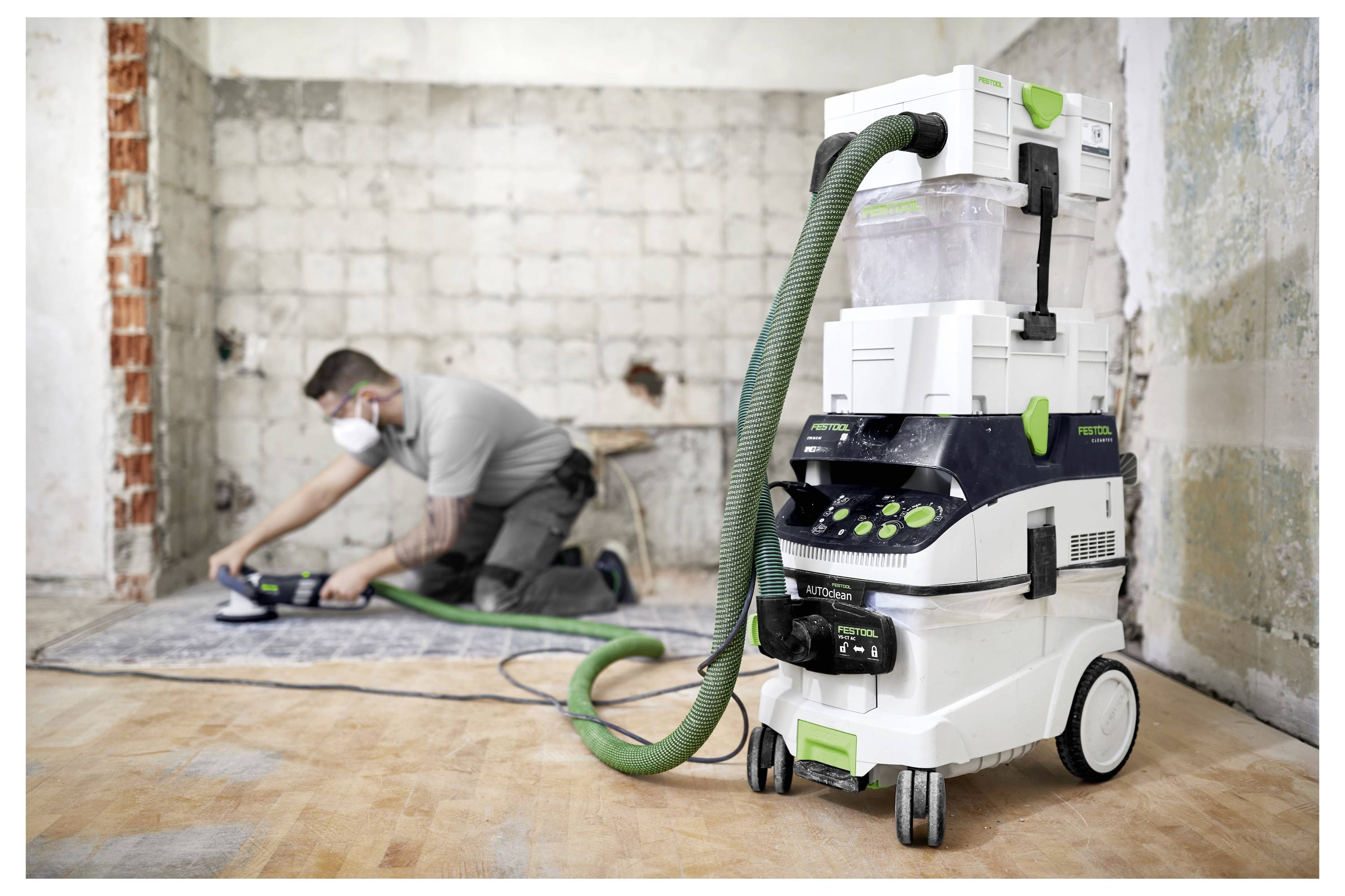 Oseba v zaščitni prašni maski dela na odstranitvi ploščic s tal. V ospredju je veliki sesalnik za ekstrakcijo prahu.