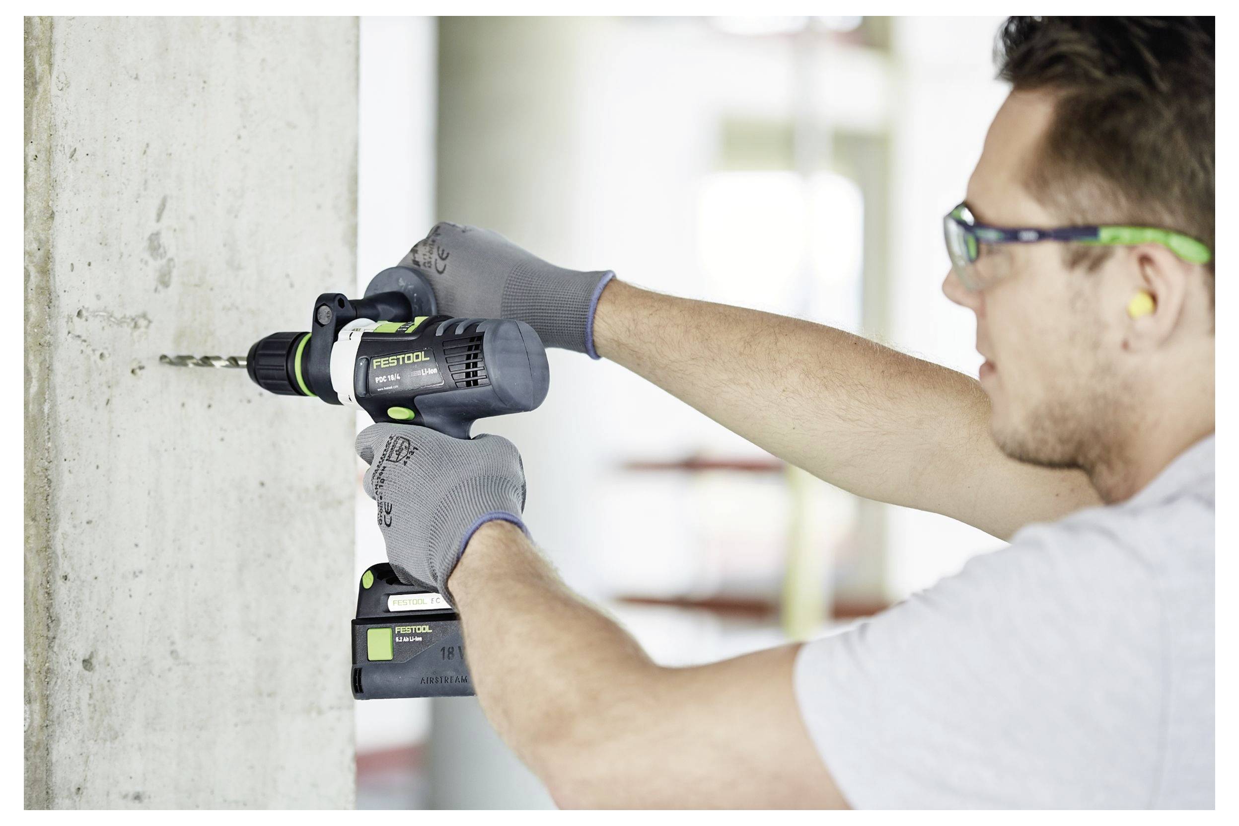Moški z zaščitnimi očali in rokavicami uporablja električno vrtalno napravo na betonski steni, kar poudarja varne gradbene prakse.