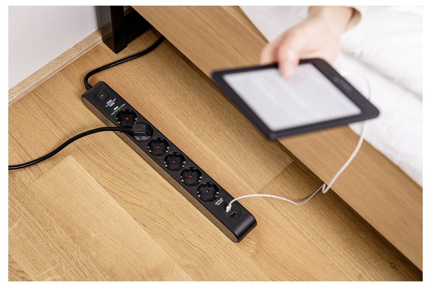 Oseba polni elektronsko napravo z uporabo USB kabla, povezanega na razdelilnik toka na lesenem podu poleg postelje.