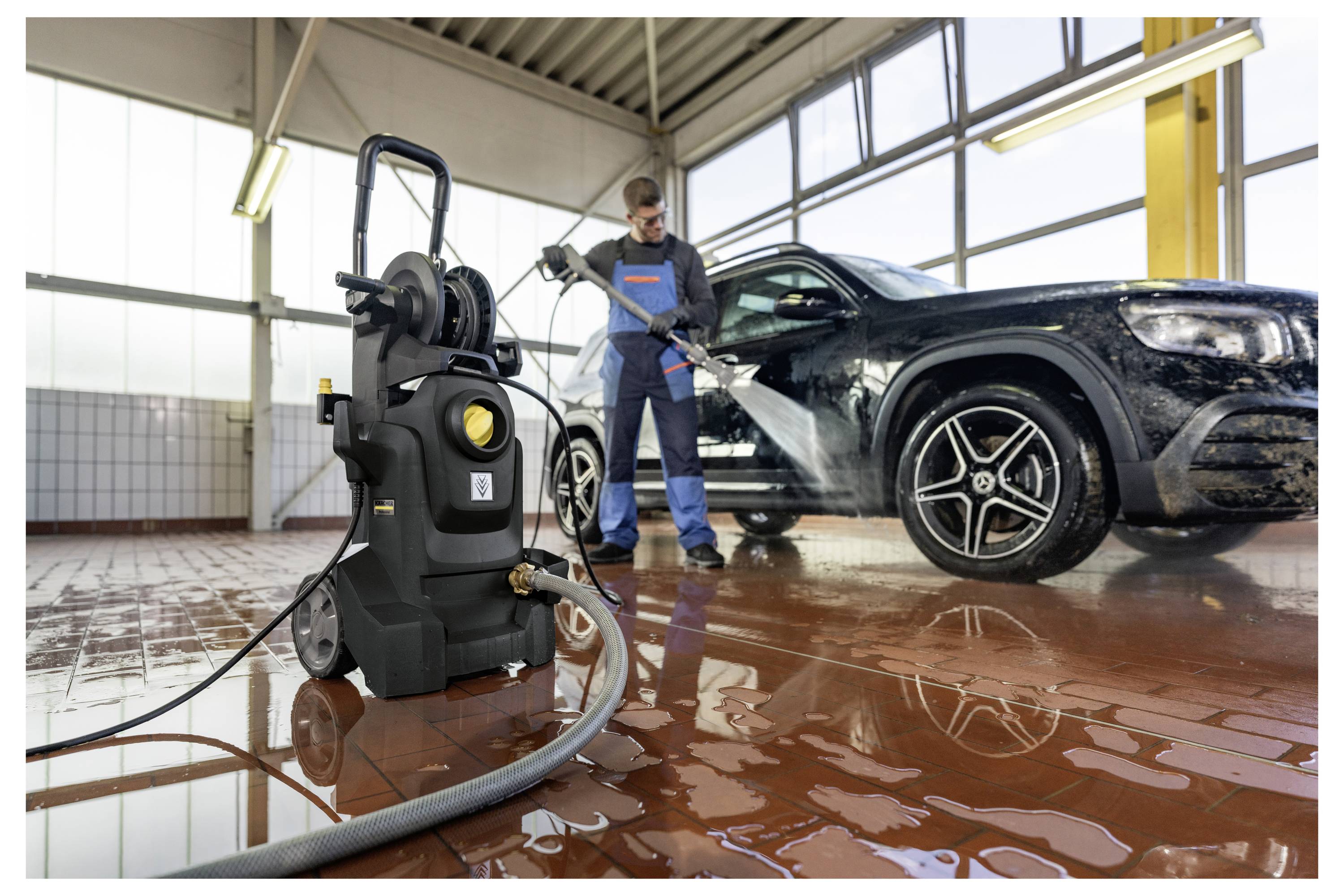 Oseba v modrih delovnih oblekah uporablja tlačni čistilnik za čiščenje črnega avtomobila v svetli garaži s ploščicami na podlagi in velikimi okni.
