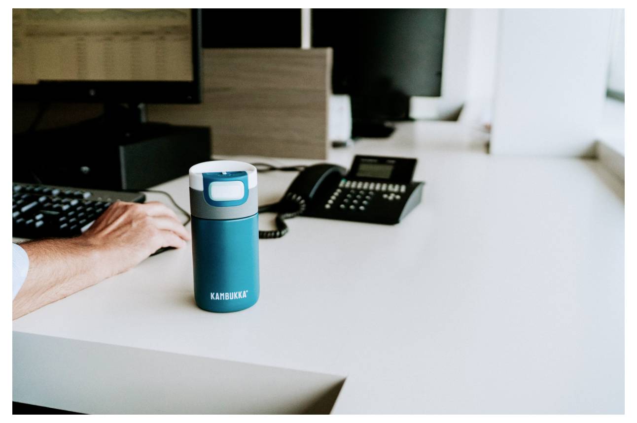 Oseba, ki uporablja tipkovnico ob modri potovalni skodelici na belem pisarnem mizi, s telefonom in monitorji v ozadju.