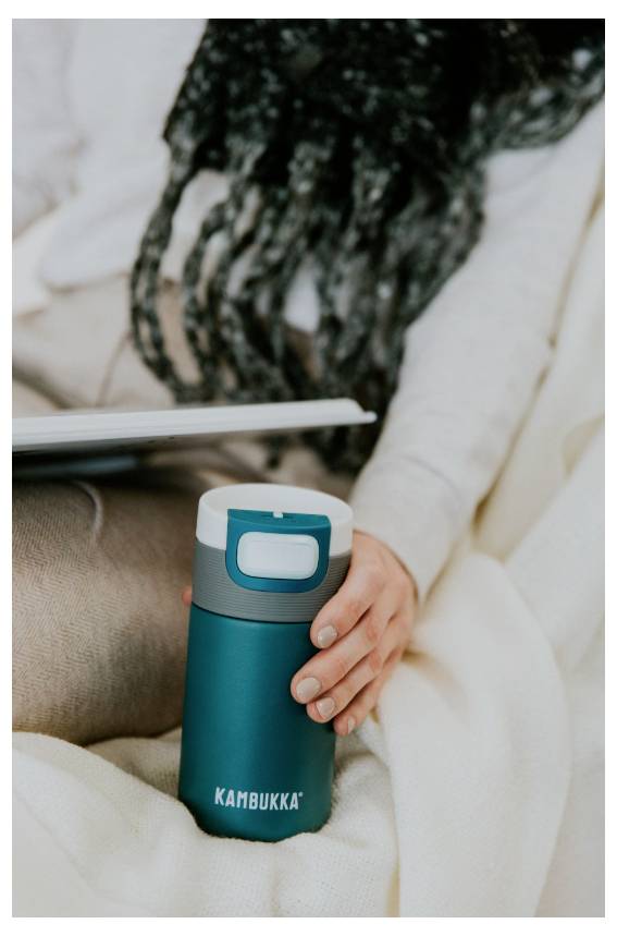Oseba sedi na kavču in drži tyrkizno toplotno izoliran lonec z napisom »KAMBUKKA«, na njenih kolenih pa je prenosnik.