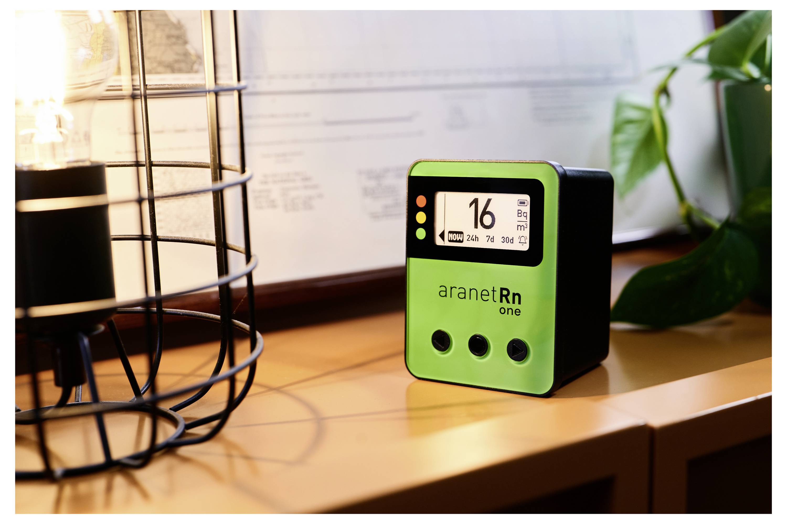Zeleni merilnik radona na mizi prikazuje '16 Bq/m³'. V ozadju sta vidna luč in rastlina.
