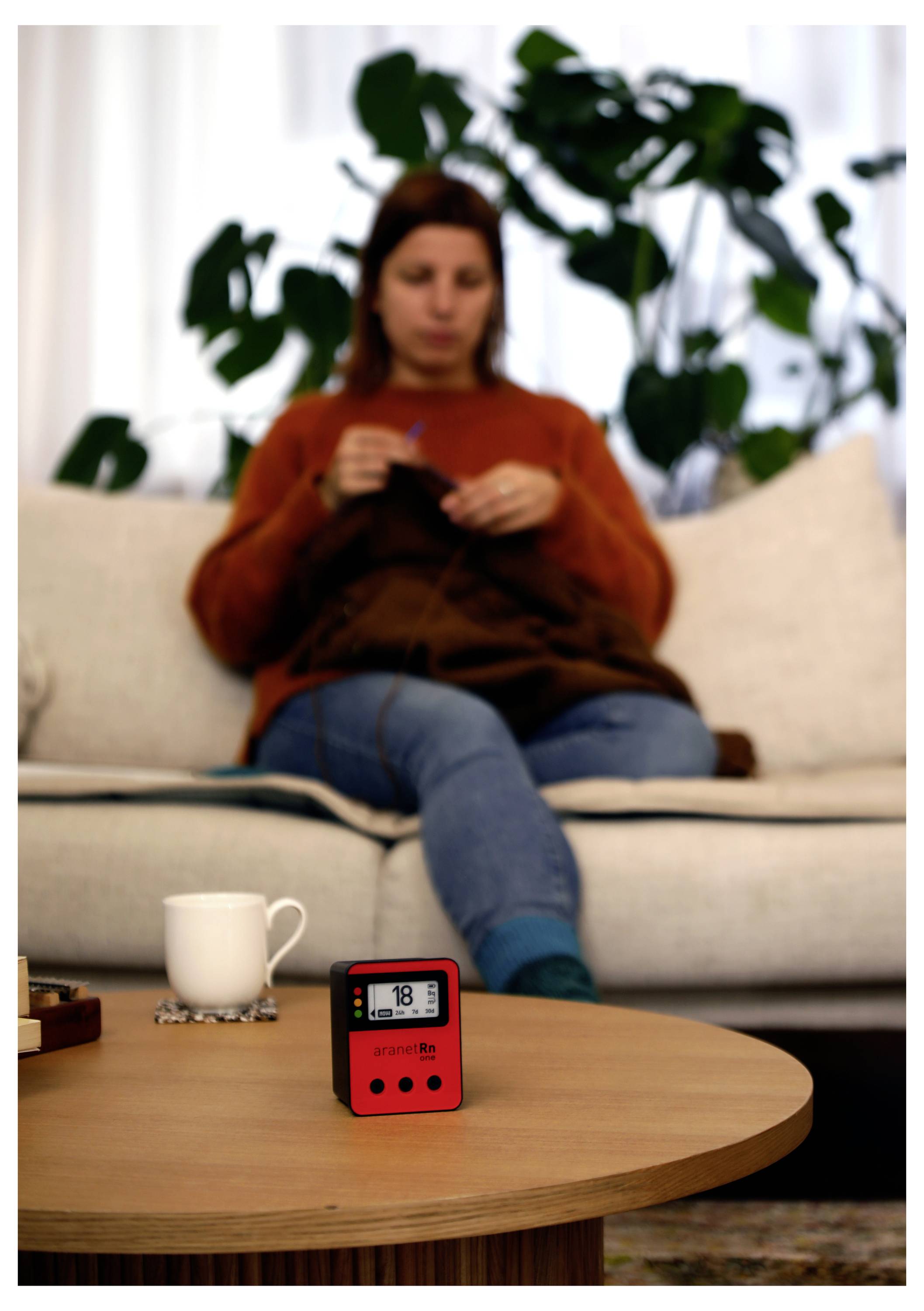 Žena sedi na kavču v ozadju in pleče. V ospredju je na kavčni mizi rdeč digitalni časovnik poleg skodelice.