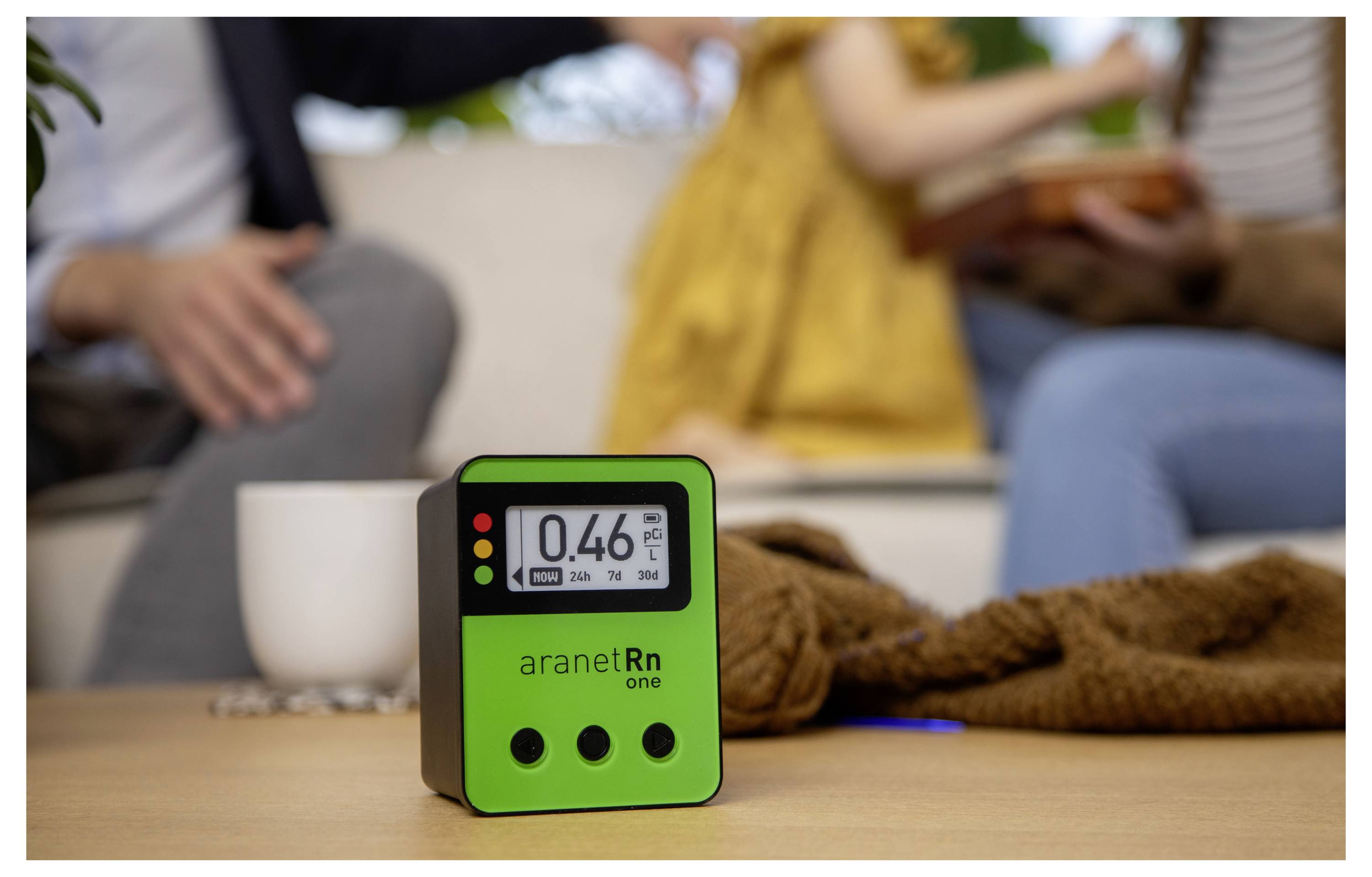 Merilnik radona z zaslonom »0,46« v ospredju, zamegljeni ljudje v ozadju, nakazuje na kakovost zraka v stanovanjskih prostorih.