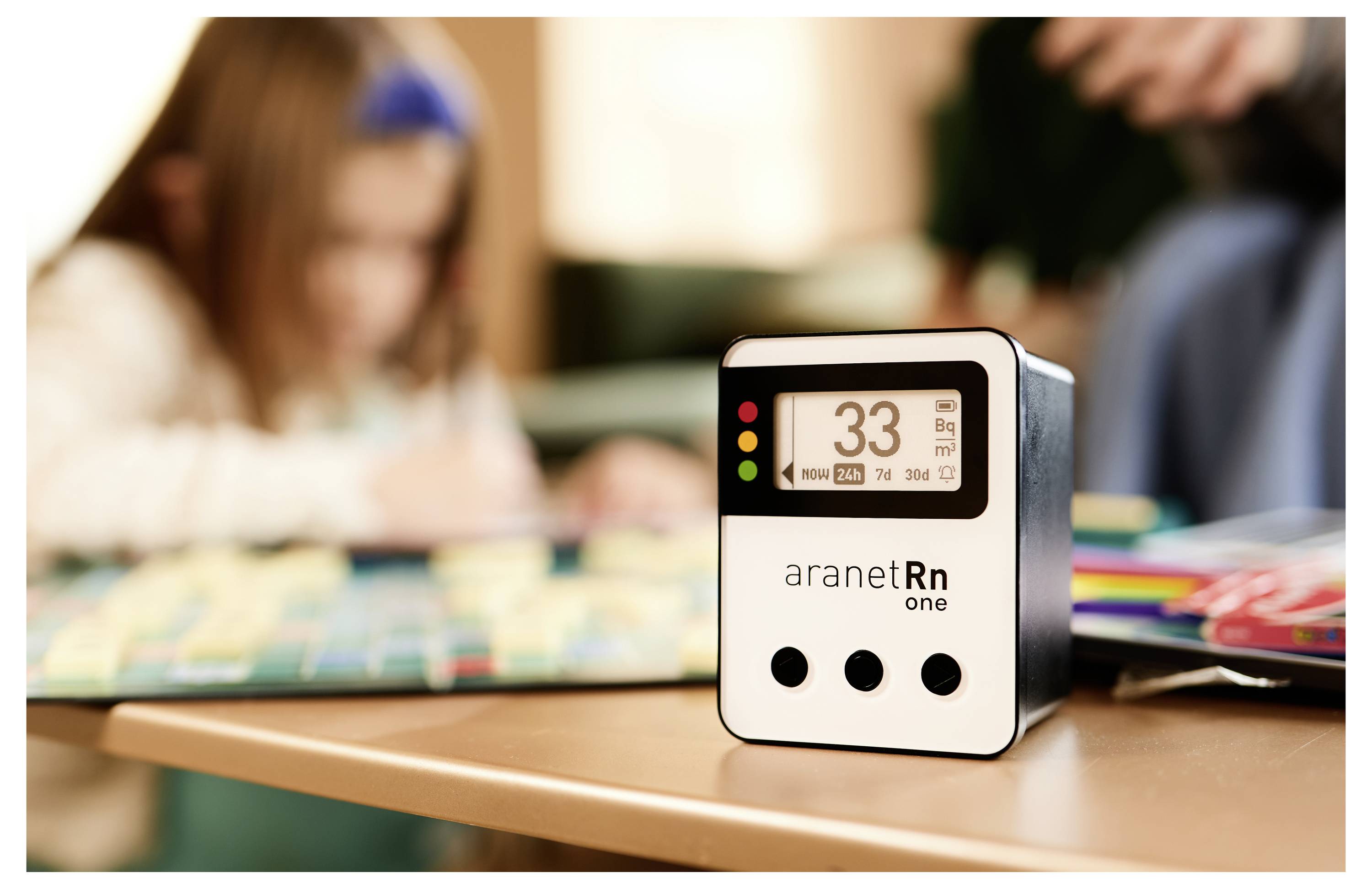 Naprava Aranet Rn One prikazuje meritev radona 33. V ozadju je otrok, ki se pravi v notranjem prostoru.