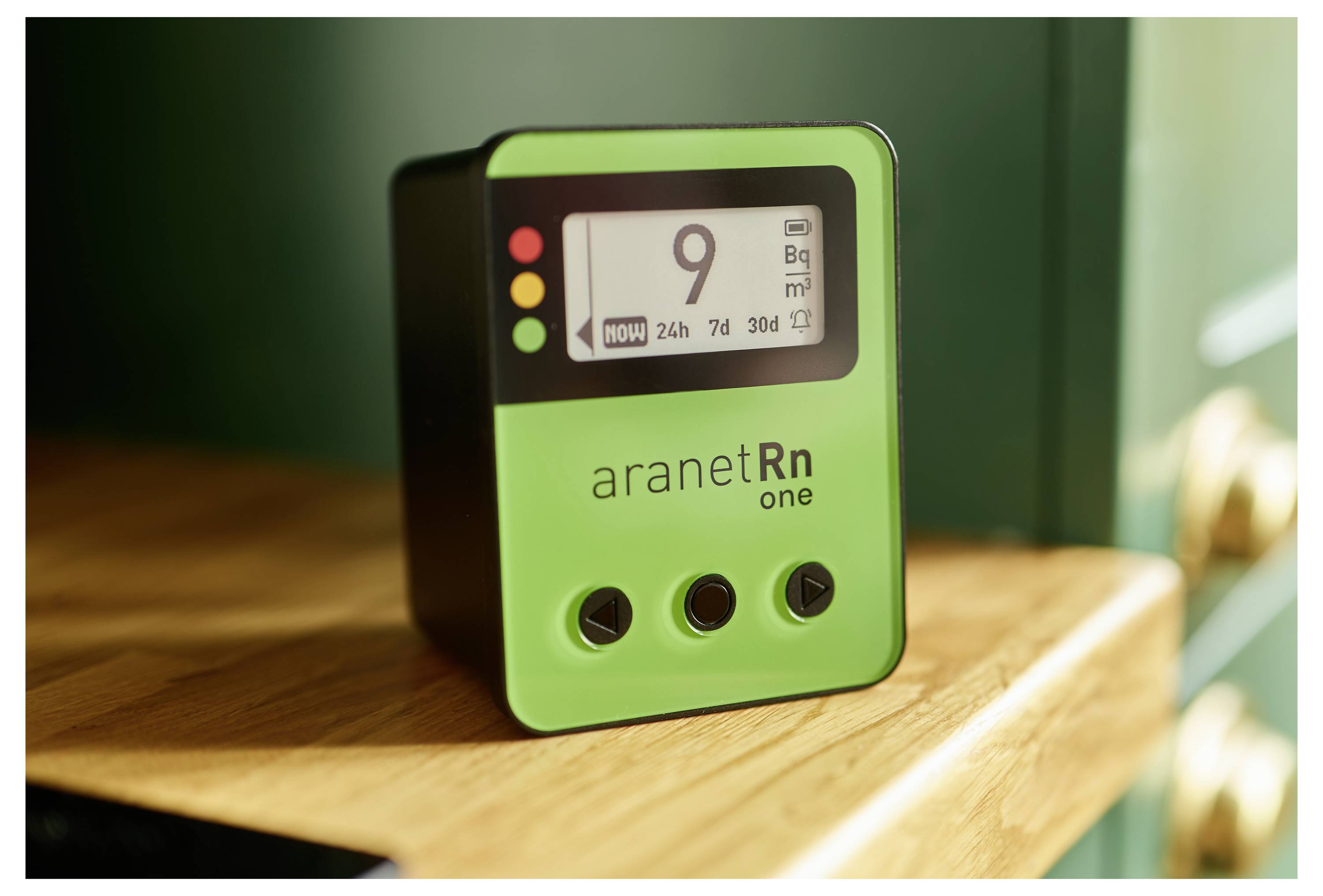 Zelena naprava prikazuje koncentracijo radona na zaslonu. Stoji na lesenem polici in ima tri krmilne gumbe.