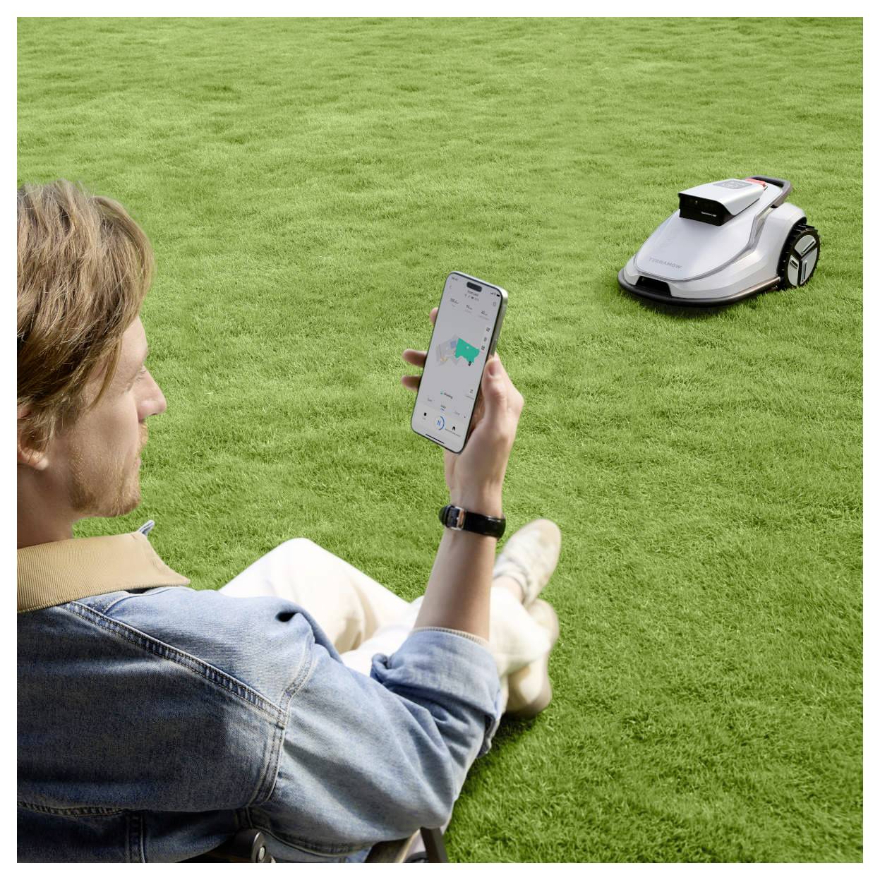Oseba na travi upravlja robotski rezalnik za travo s pomočjo aplikacije na pametnem telefonu, kar kaže povezanost in tehnologijo daljinskega upravljanja v vrtnarstvu.