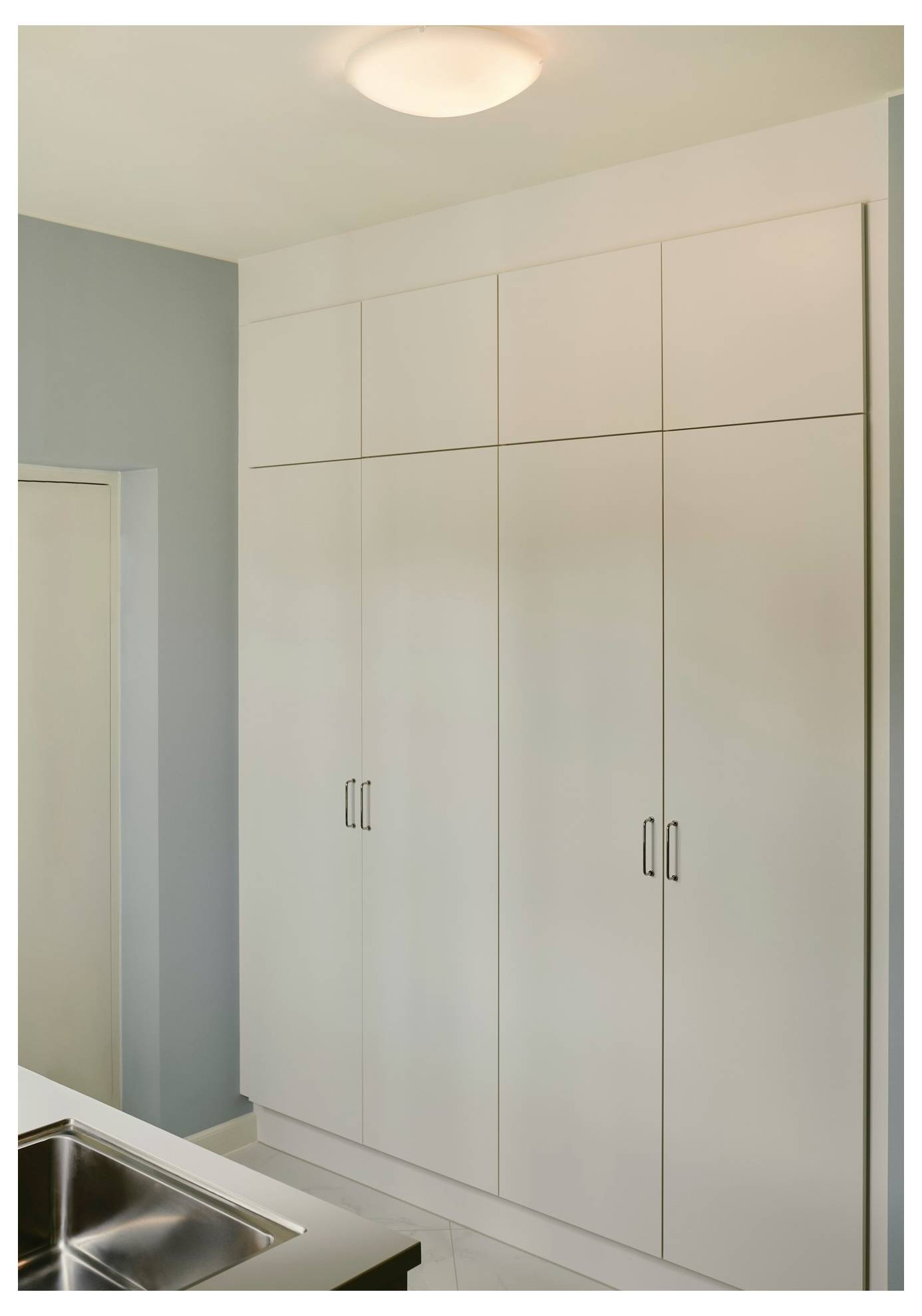 Visoki beli kuhinjski omari s stalinastimi kovinskimi ročaji, postavljeni ob svetlo modri steni, poleg nerjavečega jeklnega umivalnika.
