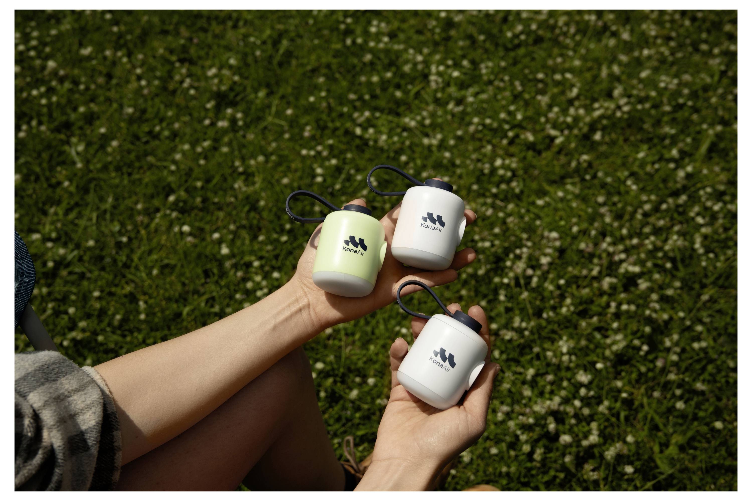 Roke držijo tri majhne vrečaste posode s zankami, dve beli in eno svetlo zeleno, pred travnatim zunanjim ozadjem.