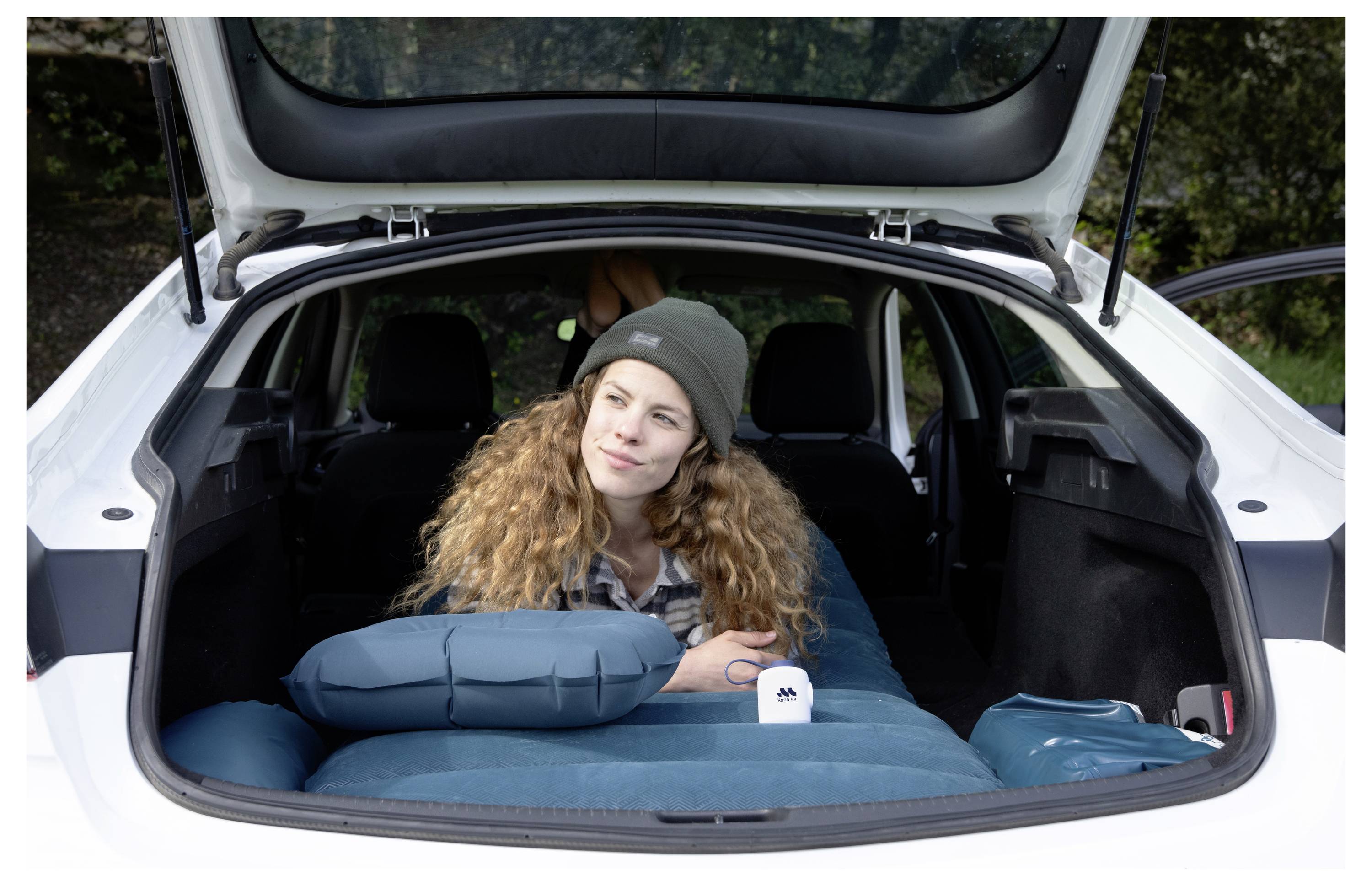 Ženska s kodrasto frizuro sedi v odprtem avtomobilskem prtljažniku, naslonjena na kampirsko opremo in blazino, kar nakazuje na avtomobilski izlet ali prestanek v naravi.