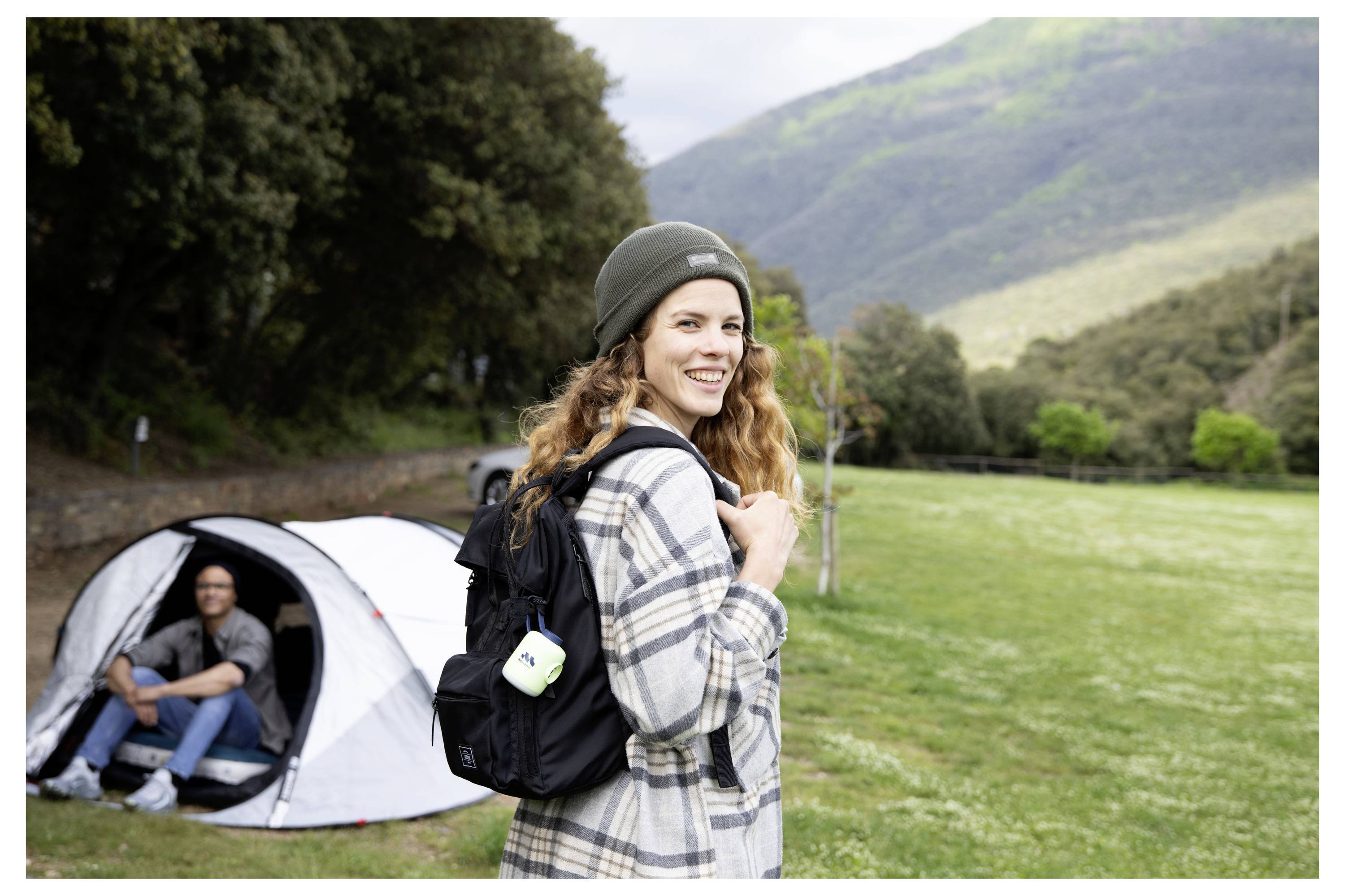 Nasmejana ženska z nahrbtnikom stoji na travnati kemping površini, moški sedi v šotoru, v ozadju so gore.