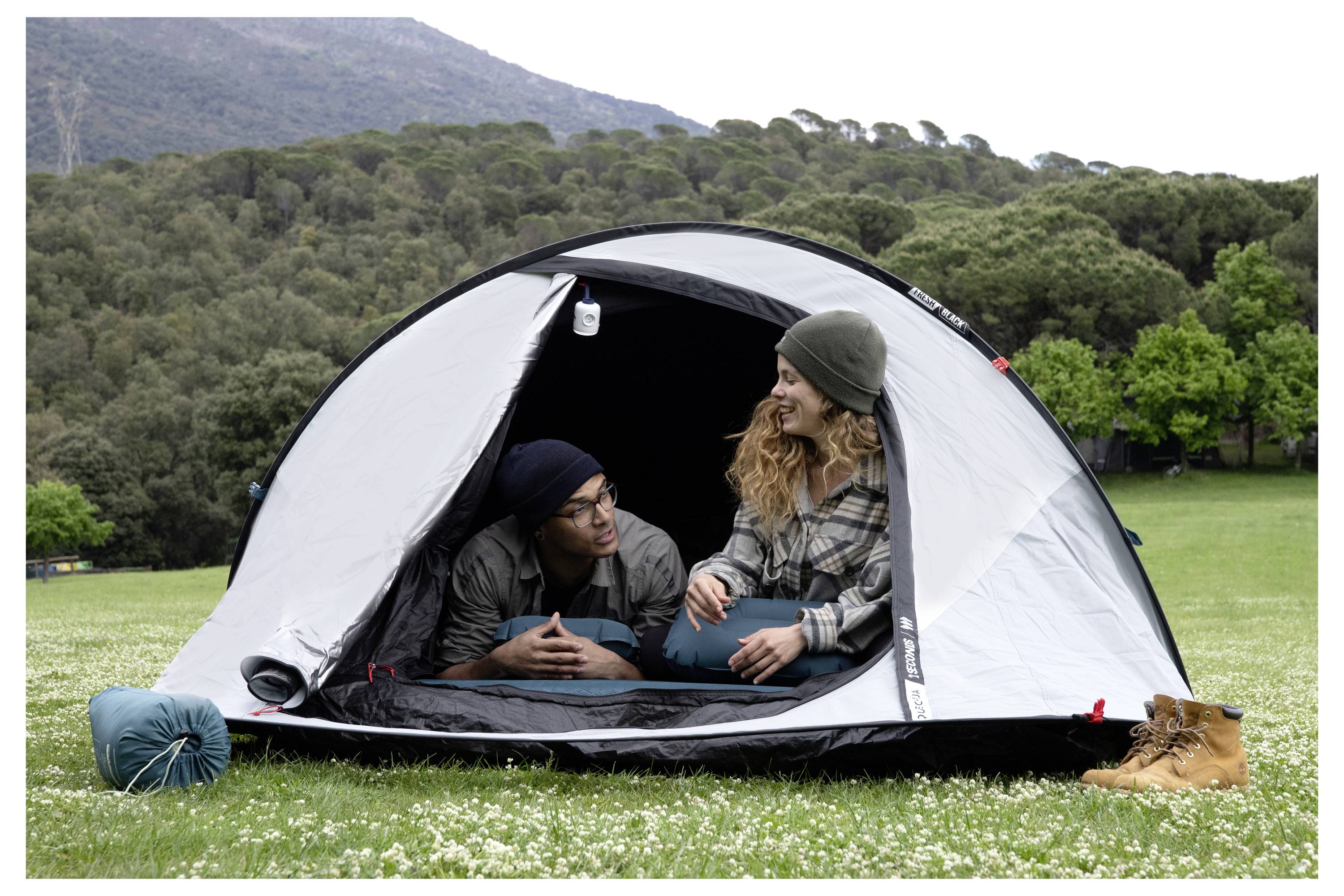 Dva kamperca sedita v sivi šotori na travniku, zunaj sta spalna vreča in čevlji, v ozadju pa se nahajajo gore in drevesa.