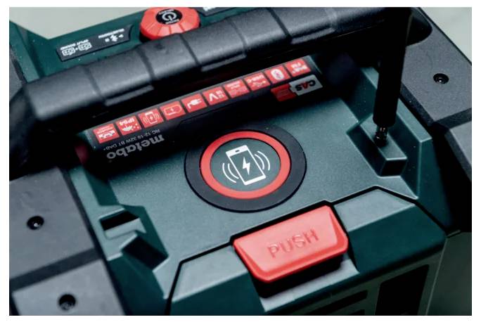 Polnilna postaja za električna orodja s rdečim gumbom »PUSH« in simbolom baterije, ki označuje funkcionalnost polnjenja. Prašnata ročka na vrhu.