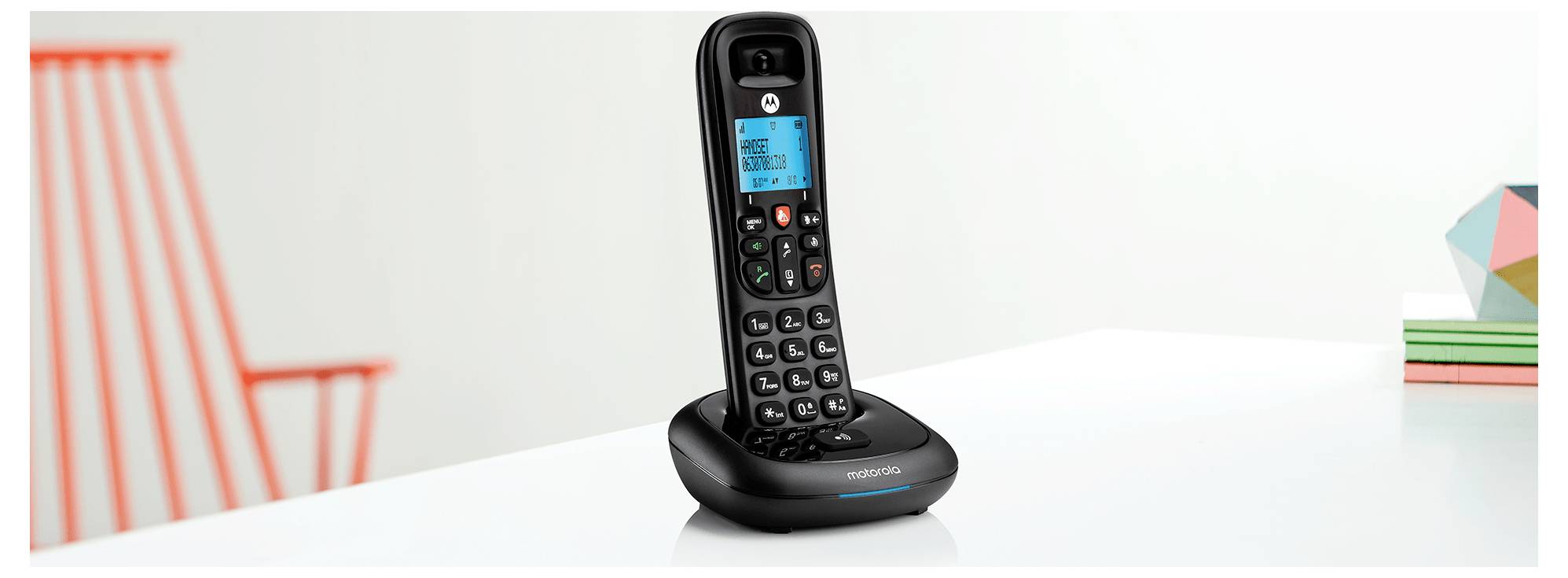 Črn brezžični telefon z digitalnim zaslonom na osnovni napravi, postavljen na beli mizi blizu barvitih geometrijskih dekorativnih predmetov.