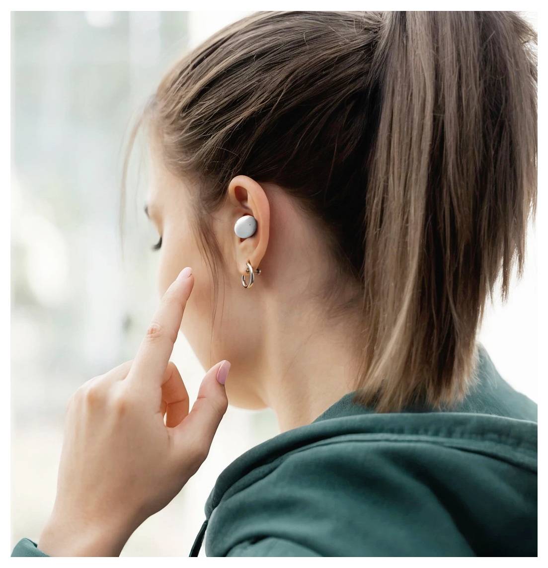 Oseba v teal puloverju, obrnjena stran, ki nosi brezžično slušalko in obročasto uhano, se dotika uha.