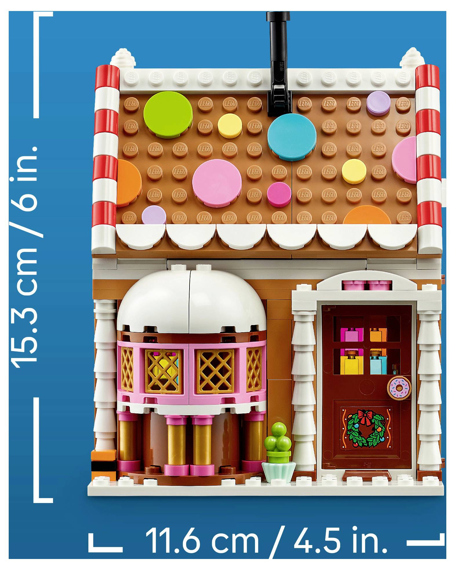LEGO-slogov miniaturni sladkarni hišica s pisanim streho iz sladkornih kapljic, rožnatimi okni, vhodnimi vrati in označbami meritev, ki prikazujejo višino 15,3 cm in širino 11,6 cm.