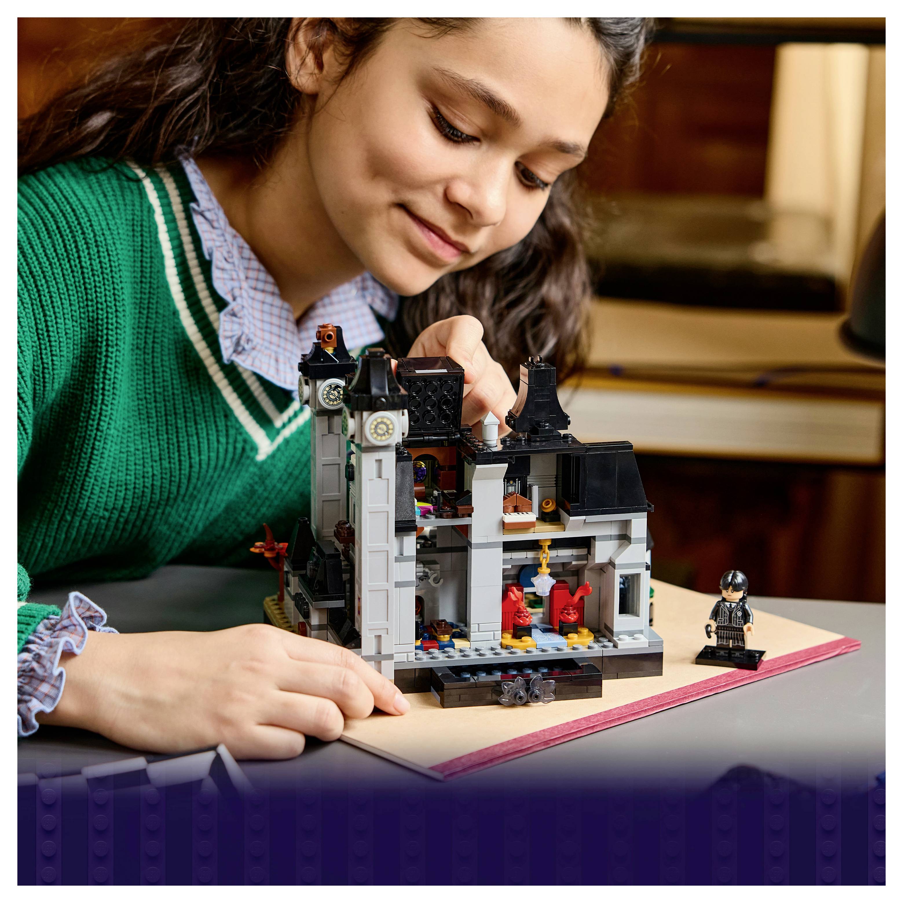 Deklica v zelenem puloverju gradi podrobno LEGO hišo na zvezku, osredotočena na model s figurico poleg njega.