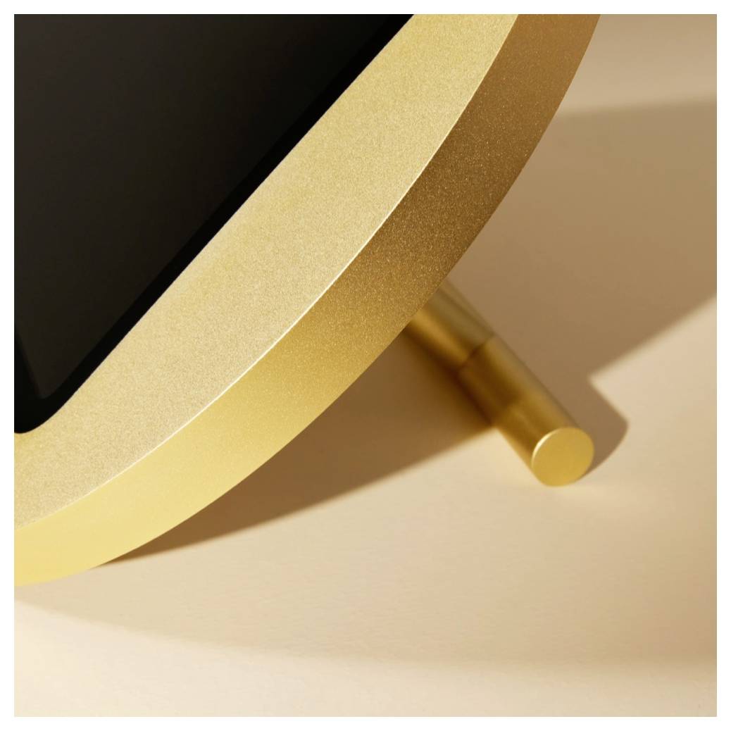Detaljni pogled na elektronsko napravo z zlatim robom, črnim zaslonom in kovinskim stojalom ali tečajem na beige površini.